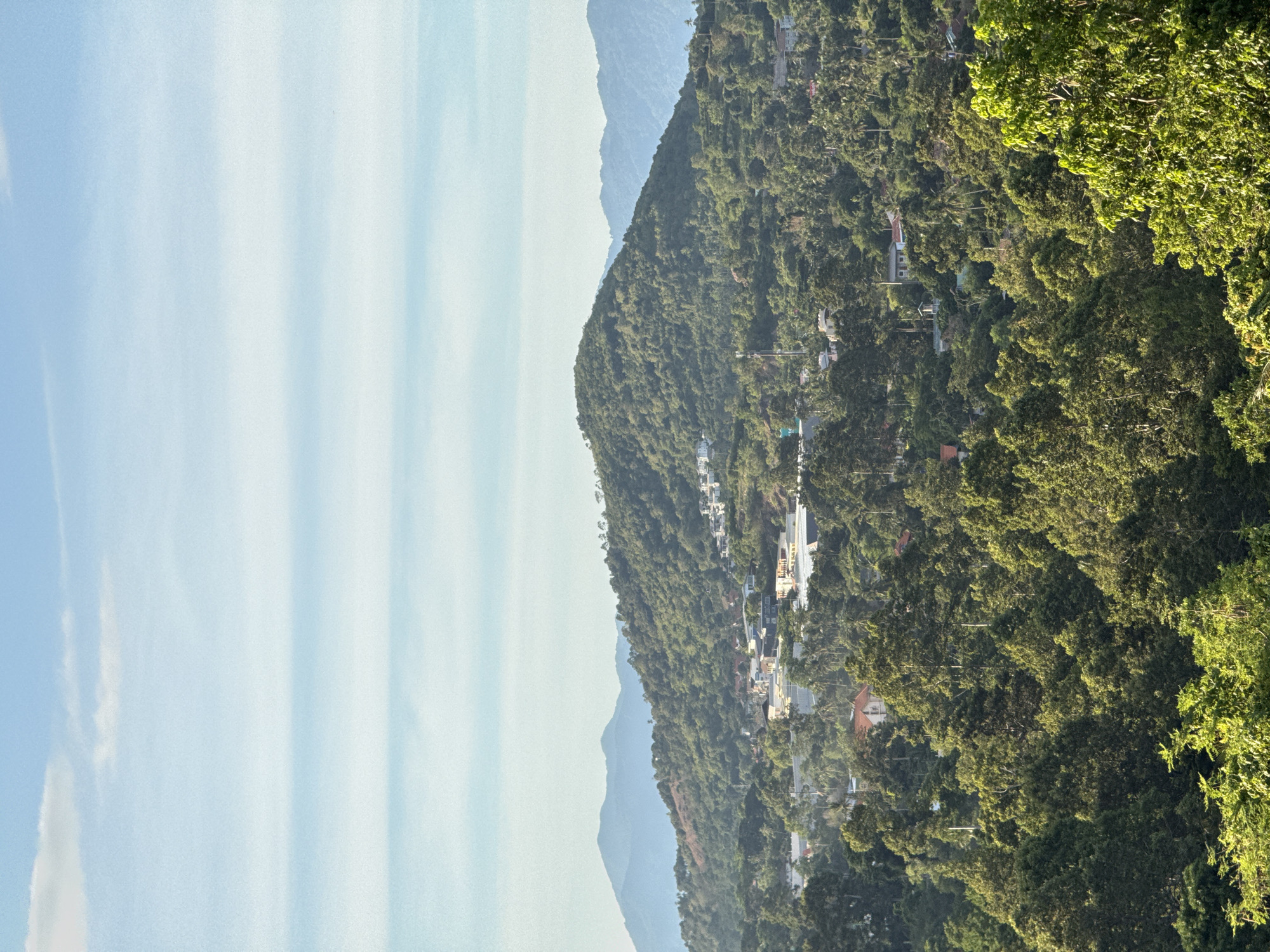 A high-angle, eye-level, long shot captures a lush, green mountainous landscape with a scattering of buildings and a bright blue sky with thin, horizontal clouds. The scene appears to be Baan Chaweng, Thailand. The foreground is dominated by dense, verdant trees, their leaves illuminated by sunlight, creating dappled shadows. Beyond this, the midground reveals a rolling green hill covered in trees, with clusters of white and beige buildings, some with red roofs, nestled amongst them. A few structures appear to be modern villas. In the background, several layers of hazy, blue mountains recede into the distance. The sky is a clear, pale blue, streaked with horizontal bands of lighter blue and white clouds. There are no people or animals visible in the image.