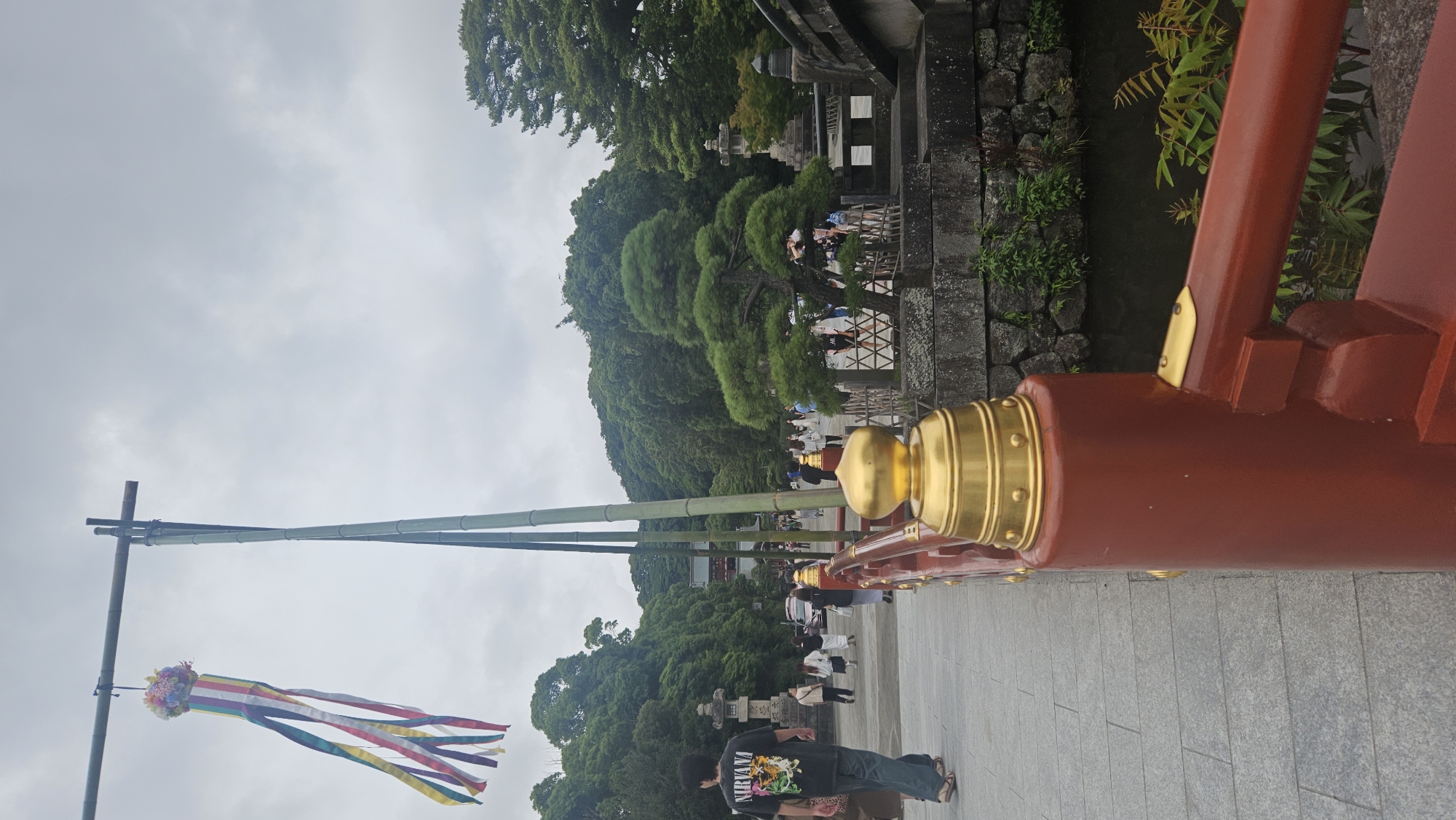 The image depicts a view of the entrance bridge to Tsurugaoka Hachimangu Shrine in Kamakura, Japan. A person wearing a black t-shirt with a "NIRVANA" graphic is walking across the bridge, facing away from the camera. The bridge has red railings with gold accents. In the background, lush green trees and a cloudy sky are visible. A traditional Japanese decorative streamer, known as a "tanabata," hangs from a bamboo pole on the left side of the image. Several other people are walking on the bridge and in the background. The environment suggests a public space, likely a tourist attraction, given the presence of other visitors and the shrine architecture in the distance.