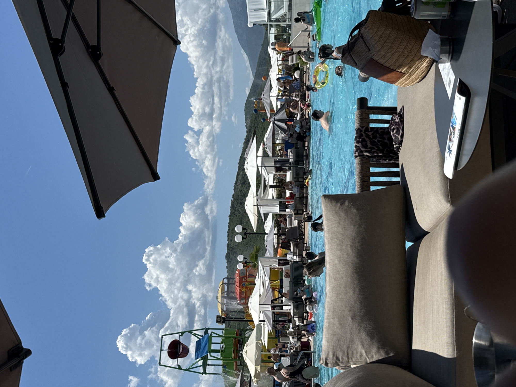 The image depicts a sunlit, outdoor scene at a resort-style swimming pool complex. In the immediate foreground, from a low vantage point, parts of a beige outdoor lounge sofa with cushions are visible, along with a dark round table to the right holding a magazine, a woven straw bag, a metal cup with a white tissue, and two beverage cans. Overhead, sections of two dark outdoor umbrellas frame the top of the image.
The midground is dominated by a large, light blue swimming pool occupied by numerous individuals, including adults and children, engaged in swimming and wading. Some children are visible with inflatable pool toys, such as a yellow ring and a green alligator. Around the pool, a multitude of white and dark sun umbrellas provide shade over lounge chairs and daybeds, some occupied by patrons. To the left, a multi-colored water play structure features a large red tipping bucket, slides, and platforms, with water actively splashing. Another smaller water feature with yellow and red elements is visible further back. Several multi-globular street lamps are interspersed throughout the pool area.
The background reveals undulating green, tree-covered hills or mountains under a bright blue sky dotted with scattered cumulus clouds, indicating a clear, sunny day. The overall scene suggests a lively leisure environment at an outdoor recreational facility in the city of Unknown, Unknown.