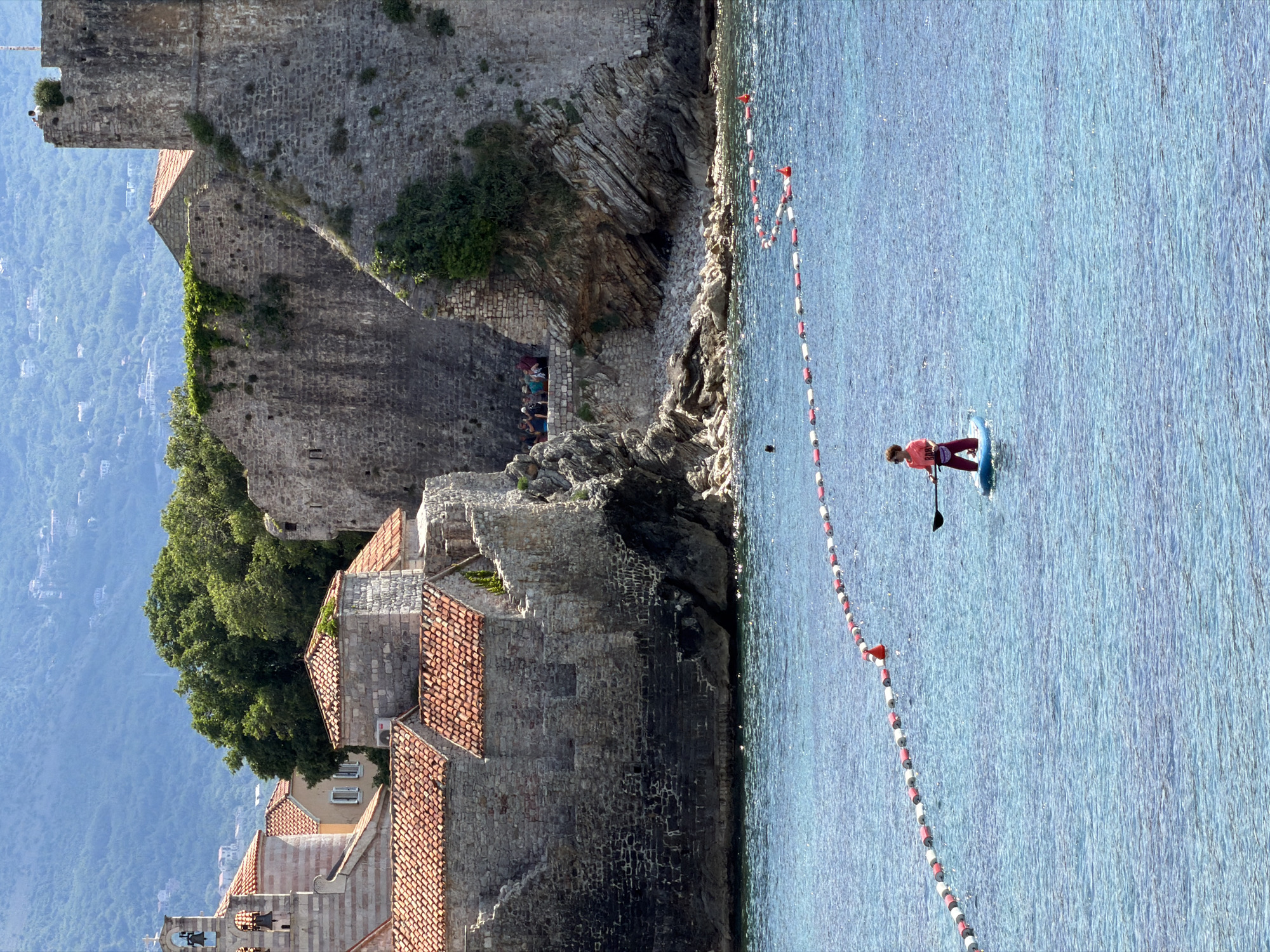 The image shows the old town of Budva, Montenegro, situated beside the Adriatic Sea. The architecture includes stone walls and buildings with red-tiled roofs, suggesting a historical or medieval style. Vegetation, including trees, is visible among the buildings. The sea is calm and light blue, with a series of red and white buoys marking a boundary. A person stands on a paddleboard, using a paddle to move through the water parallel to the old town's coastline. Several people can be seen near the base of the stone wall, presumably tourists. The backdrop includes a mountainous landscape partially obscured by haze.