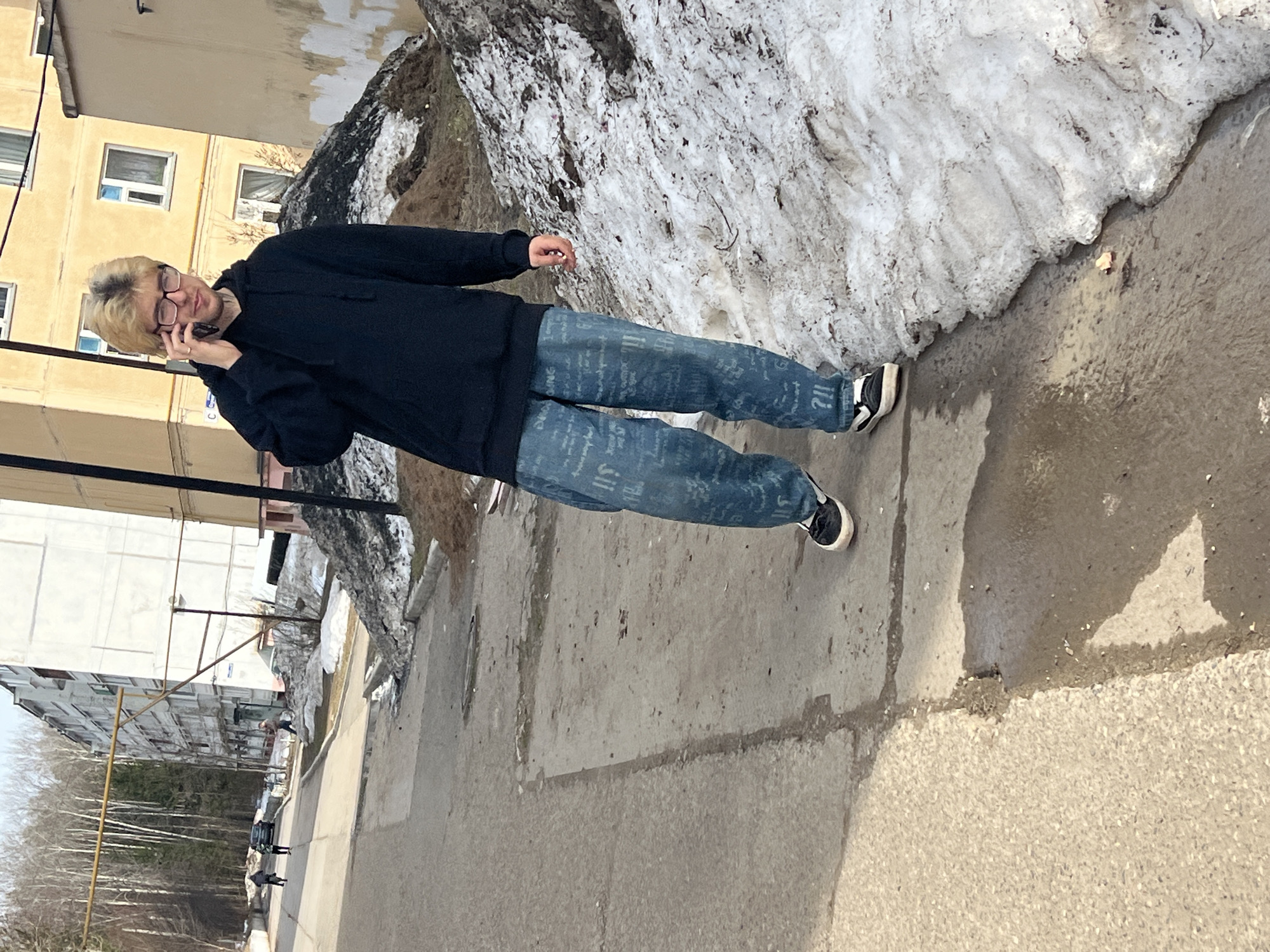 A young man is walking on a sidewalk in Noviy Urengoy, Russia while talking on his cell phone. He is wearing glasses, a dark hooded sweatshirt, blue jeans with writing on them, and black and white sneakers. The sidewalk is wet with melting snow, and there is a large pile of snow next to the sidewalk. Buildings are visible in the background.