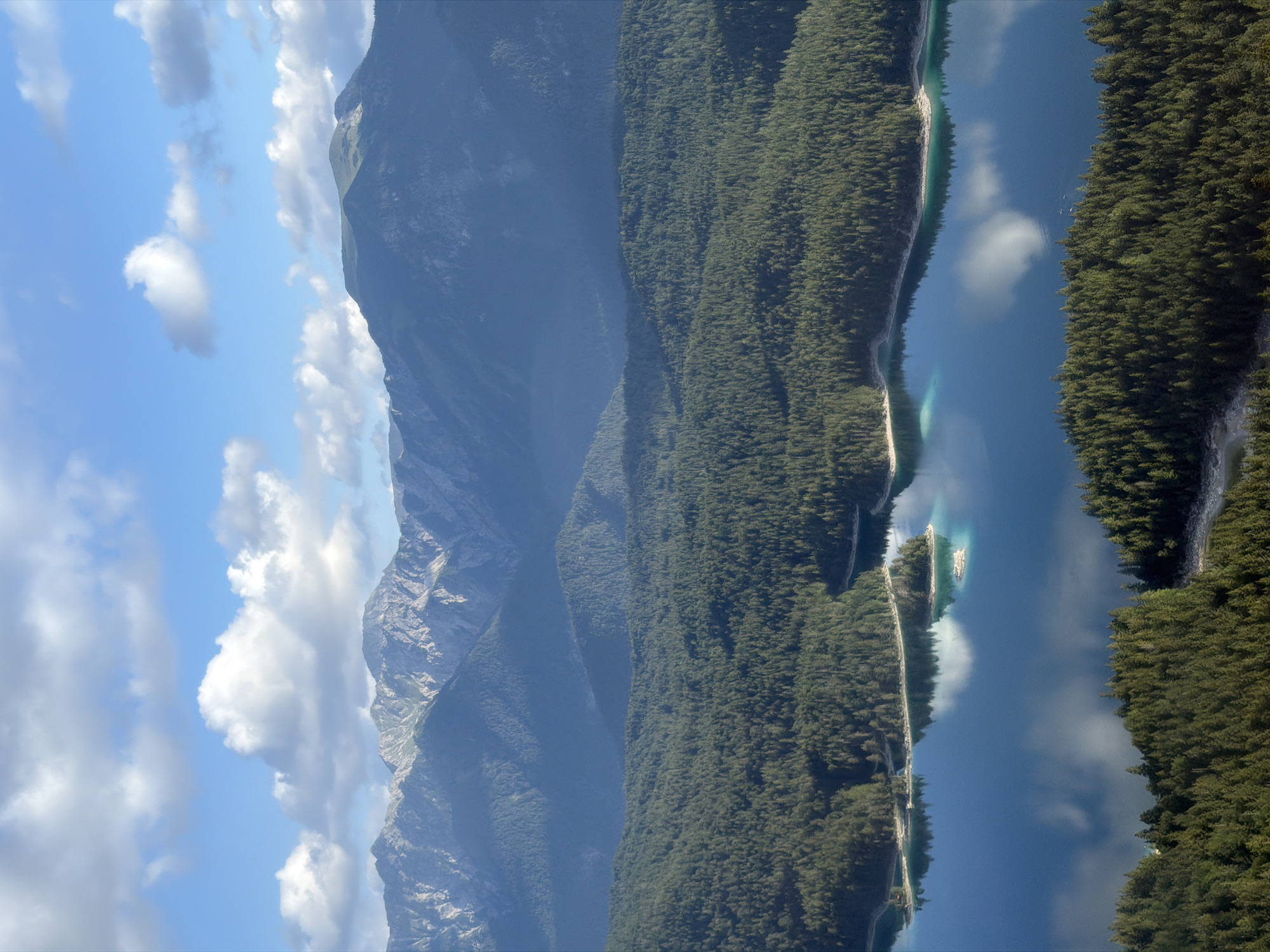 This is a high-angle, full shot of the Eibsee lake in Grainau, Germany, on a bright, partly cloudy day. In the foreground, dense, dark green coniferous trees cover the undulating hillsides surrounding the lake. The lake itself is a vibrant, deep blue, reflecting the sky and clouds. Several small, tree-covered islands dot the surface of the water. In the midground, more forested hills create a layered effect, leading the eye towards the background. The background is dominated by a range of imposing, rugged mountains with rocky, grey peaks. The sky above is a clear blue, interspersed with fluffy white clouds, some of which are casting soft reflections on the lake's surface. There are no people or animals visible in the image.