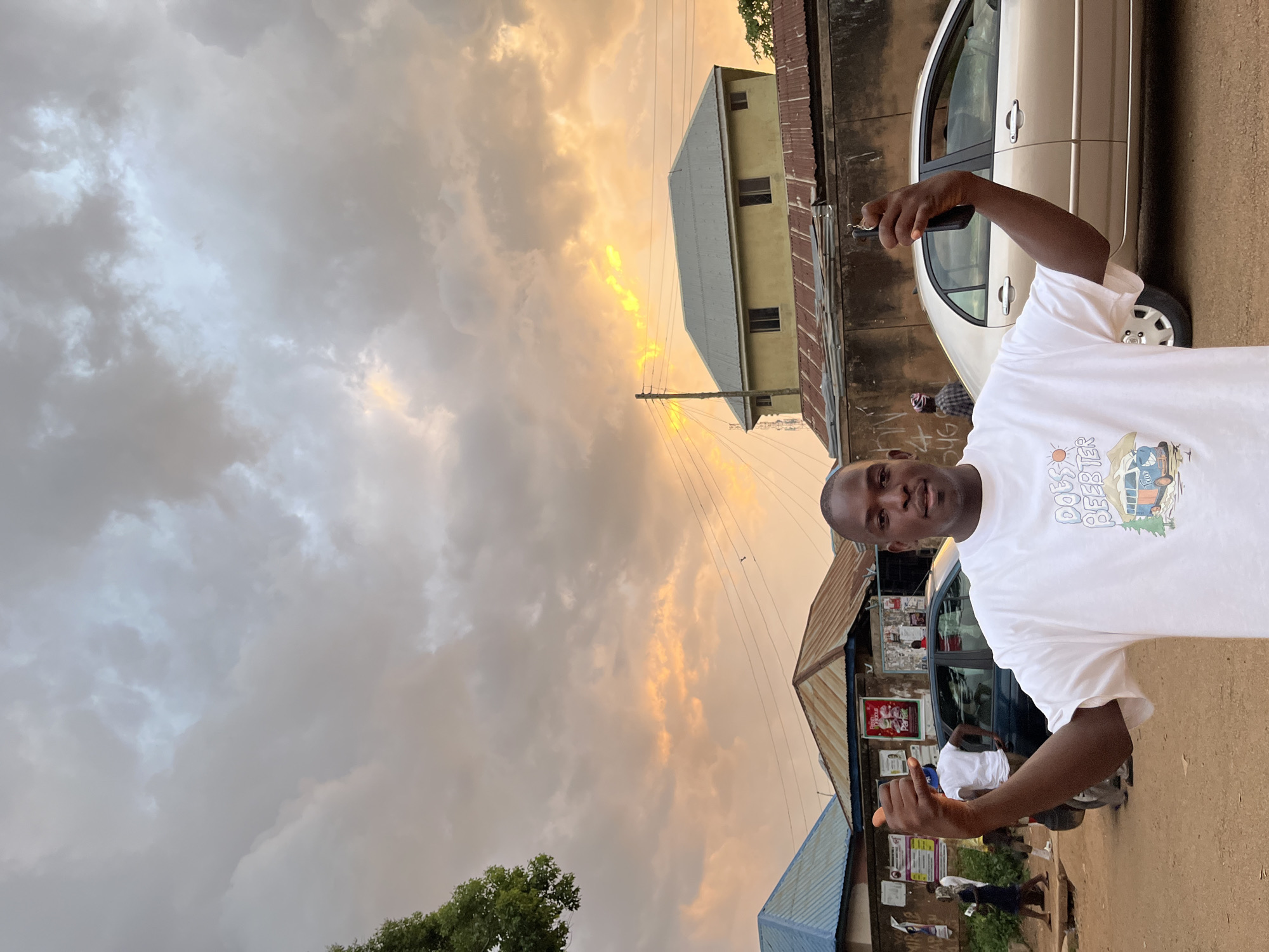 A man wearing a white t-shirt stands in the foreground with his arms raised, his left hand in a shaka sign and his right hand holding a mobile phone. His t-shirt has a graphic print of a van and text that reads "DOES BEER?". Behind him, several buildings with corrugated metal roofs line a dirt road. A light-colored sedan is parked on the right side of the frame. In the background, utility poles and wires crisscross the sky, which is filled with dramatic, cloudy formations illuminated by the warm glow of sunset, with a bright orange and yellow band visible towards the horizon. The scene appears to be an outdoor, urban environment, likely in Jos, Nigeria, given the architectural style and the presence of a street market or shops. Several other individuals are visible in the middle ground, including two men near a dark sedan and a woman walking on the left side of the road.