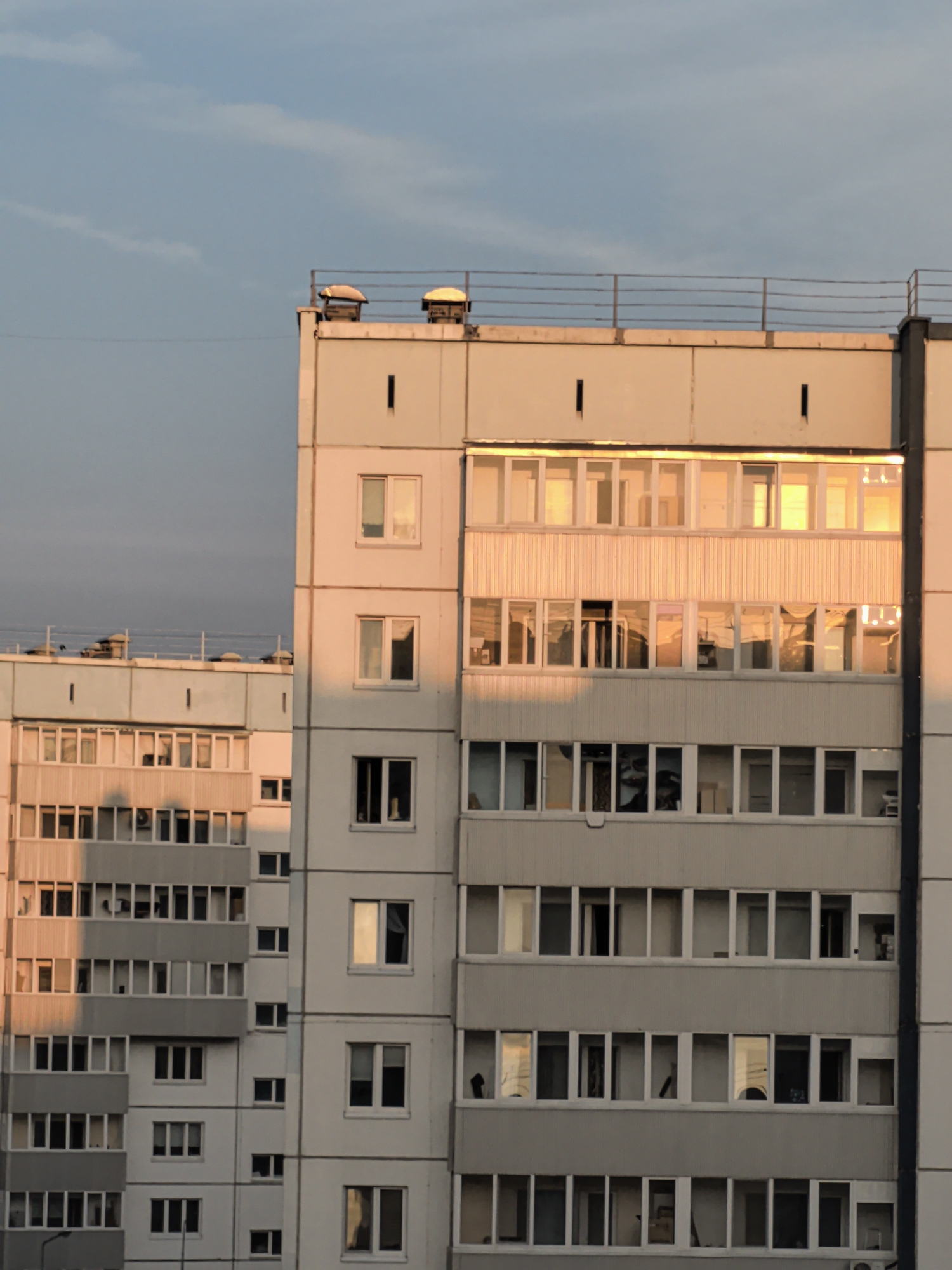 This image captures a close-up view of the upper floors of two apartment buildings in Froly, Russia, bathed in the warm, golden light of sunset. The buildings are constructed from concrete panels, typical of Soviet-era architecture. The building on the right is closer to the foreground and shows a more detailed facade. It has multiple stories, each with rows of windows, some of which are balconies. The windows on this building are mostly dark, but some reflect the sunset, showing streaks of orange and yellow. The building on the left is slightly further back and appears to have a more complex arrangement of balconies and windows. It also exhibits the same concrete panel construction and is also illuminated by the sunset. The sky above is a clear, pale blue, with faint wisps of clouds visible. The overall impression is one of a quiet urban evening scene.
