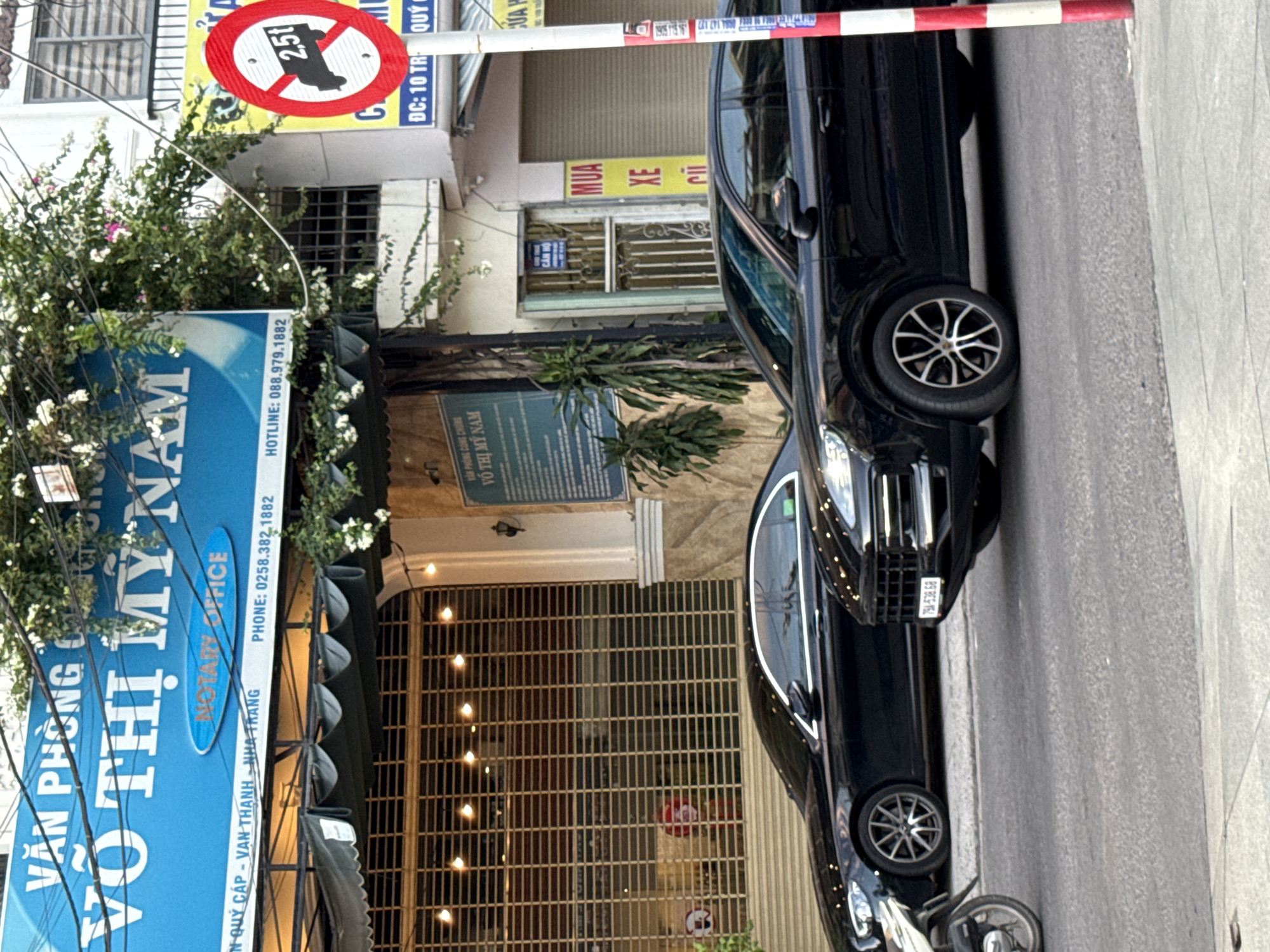 A street scene in Nha Trang, Vietnam features two black cars parked or driving on a paved road. In the background, a building displays a blue sign with Vietnamese text and "NOTARY OFFICE" in English. Further to the right, a red and white striped pole stands beside a circular traffic sign prohibiting vehicles over 2.5 tons. The street is lined with shops, some with metal roll-down shutters. Foliage and flowering plants are visible on and around the buildings. The overall impression is that of a commercial street in an urban environment.