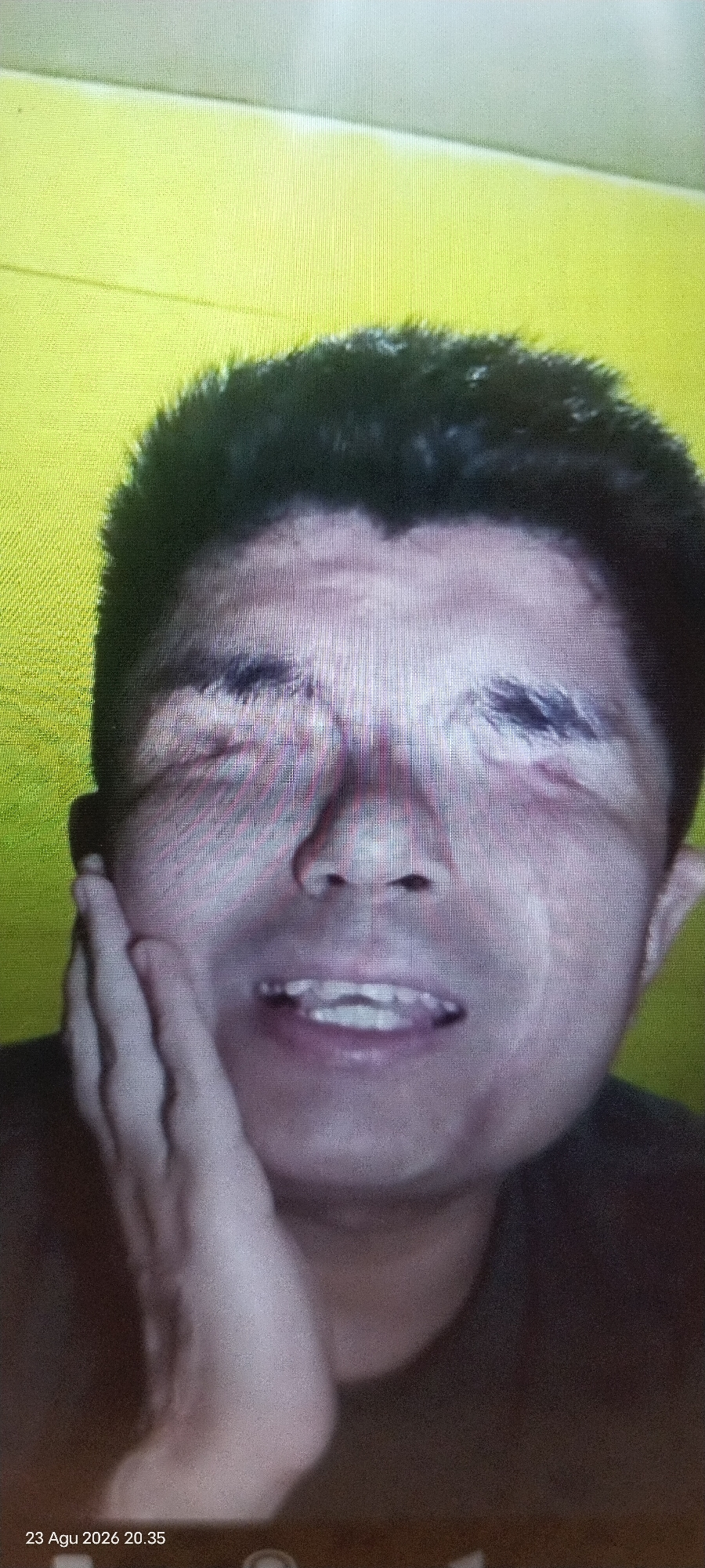A close-up, low-resolution, digitally distorted selfie shows a person's face. The subject's eyes are closed, and their mouth is open, revealing teeth. Their right hand is touching their cheek. The background is a bright, uniform yellow. The image is timestamped "23 Agu 2026 20.35". The location is East Jakarta, Indonesia.