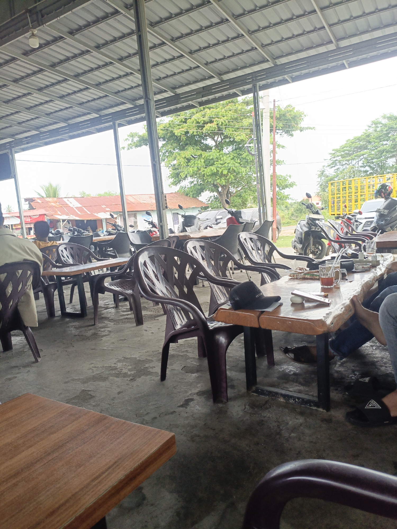 The image depicts an outdoor seating area at what appears to be a roadside eatery in Lhokseumawe, Indonesia. The scene is covered by a corrugated metal roof supported by metal beams, suggesting an open-air structure. In the foreground, several plastic chairs surround wooden tables. On one of the tables, there is a glass of iced tea, a small plate of food, a black baseball cap, and a mobile phone. The ground is concrete, showing signs of wear and tear. In the background, several motorcycles are parked, indicating that customers likely arrived by motorbike. Beyond the seating area, there are buildings with red roofs and trees, suggesting a semi-urban or rural environment. The lighting is overcast, consistent with a cloudy day. The overall impression is of a casual dining establishment catering to motorcyclists and locals in Lhokseumawe.