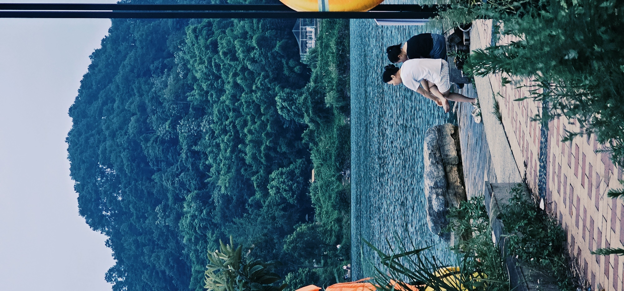 In Chongqing High-tech Zone, China, two women are seated on a stone bench by a body of water. The woman on the left wears a white t-shirt and checkered shorts, while the woman on the right is dressed in a black t-shirt and dark shorts. They are positioned facing away from the camera, looking out at the water. A large rock is situated to their left. The foreground features a tiled walkway with green foliage. Behind the water, a lush, tree-covered hillside rises, culminating in a clear sky.