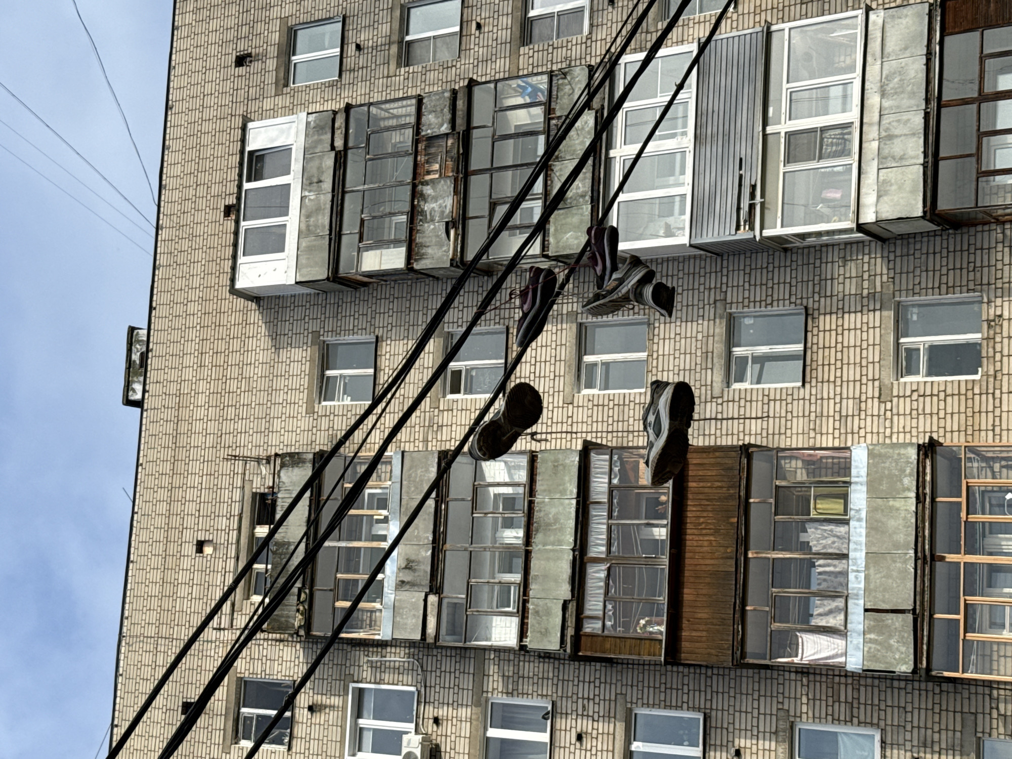 A bright, daylight scene in Rusanovo, Russia, showcases a segment of a multi-story apartment building against a clear blue sky. The building features a facade constructed from small, light-colored bricks. Its windows exhibit a variety of styles; some are standard, while others are enclosed as small, ad-hoc balconies or sunrooms, utilizing diverse materials such as glass, corrugated metal, and wooden panels, suggesting individual modifications.
Several dark utility cables run diagonally across the foreground, from the upper left to the lower right. From these cables, multiple pairs of shoes, specifically sneakers or trainers, are suspended by their laces. At least three distinct pairs are visible, generally dark in color with lighter soles. This act of "shoe-tossing" is a prominent and unusual detail in the urban environment.
The time of day is clearly bright and sunny, indicated by the well-lit building and the cloudless sky. No people are visible, and no ongoing activity is captured, only the static result of the shoes being hung on the wires. The utilitarian architecture with its varied, individually enclosed extensions serves as a contextual cue for its location in a post-Soviet urban setting. A small, white air conditioning unit is visible on the lower part of the building. There is no visible text in the image.