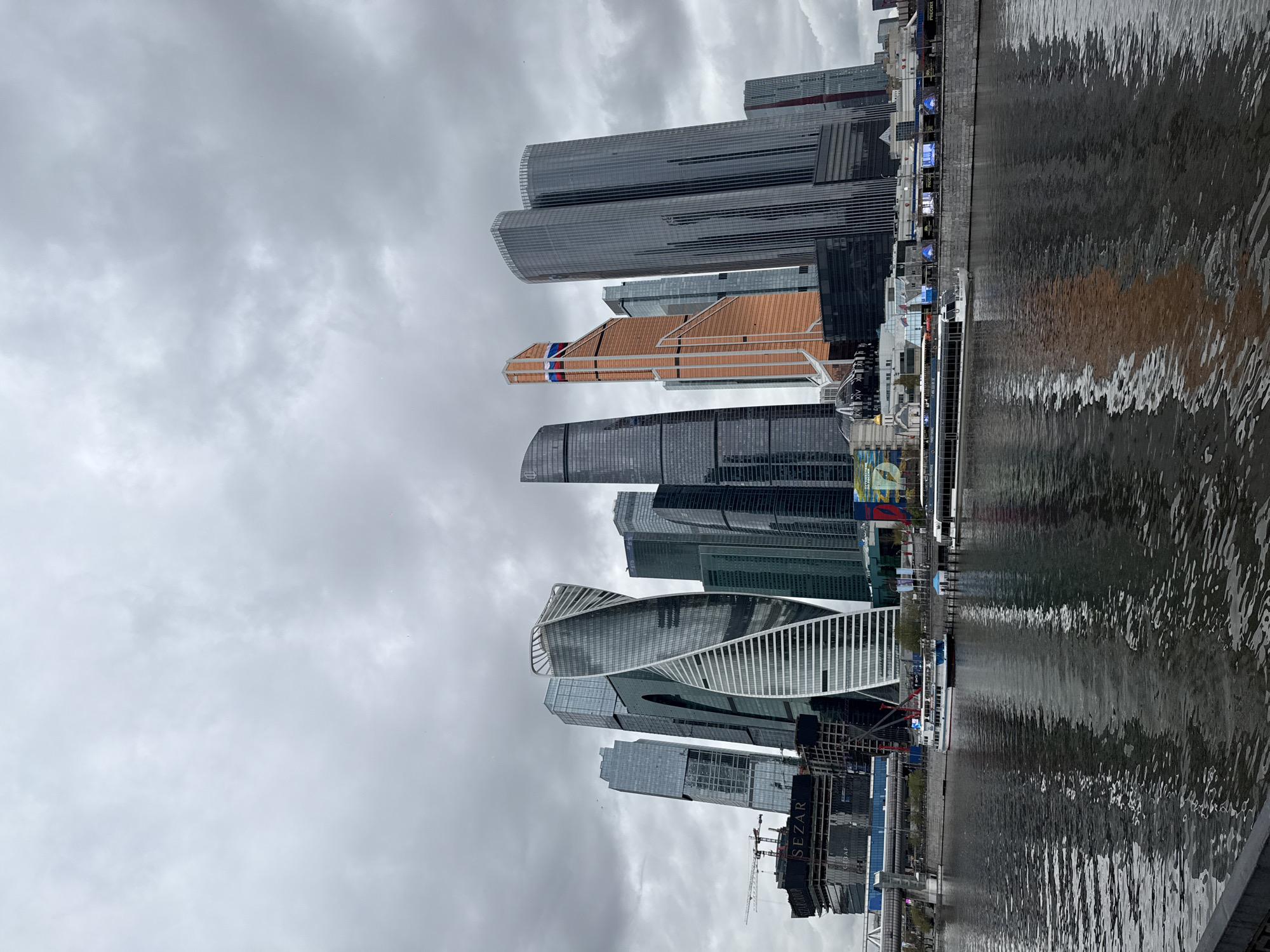 The image depicts the Moscow International Business Center ("Moscow City") reflected in the Moscow River. The skyline includes several modern skyscrapers of varying heights and designs, constructed primarily of glass and steel. Dominant structures feature a mix of rectangular, cylindrical, and uniquely twisted architectural shapes. The image is taken under a cloudy, overcast sky. The river reflects the cityscape, albeit with distortion caused by ripples. A boat is docked along the riverbank. Construction activity is visible, including cranes and ongoing building projects.