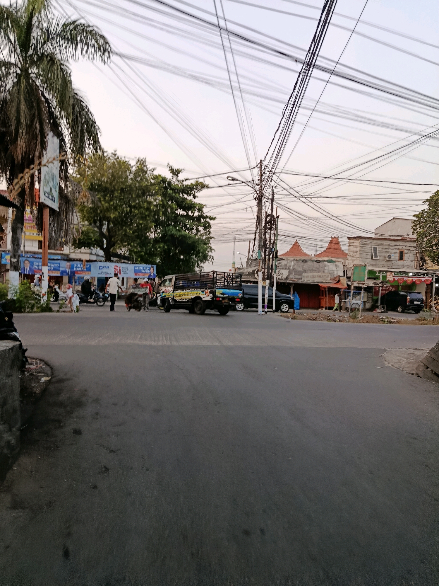 The image depicts a street scene in Bekasi, Indonesia, with a focus on a busy intersection. Power lines crisscross the sky, forming a dense network above the street. In the foreground, a gray asphalt road is visible, with some patches of discoloration and debris.
On the left side of the road, a tall palm tree stands next to a building adorned with posters and banners, some featuring images of politicians. Several people are present, with some on motorcycles. Further down the street, a pickup truck with a cage-like bed is parked.
The background reveals a collection of buildings, some with distinct tiled roofs, suggesting a mix of commercial and residential structures. A few people are seen walking in front of these buildings, and another dark-colored vehicle is parked. The overall atmosphere suggests a typical day in a bustling urban environment.