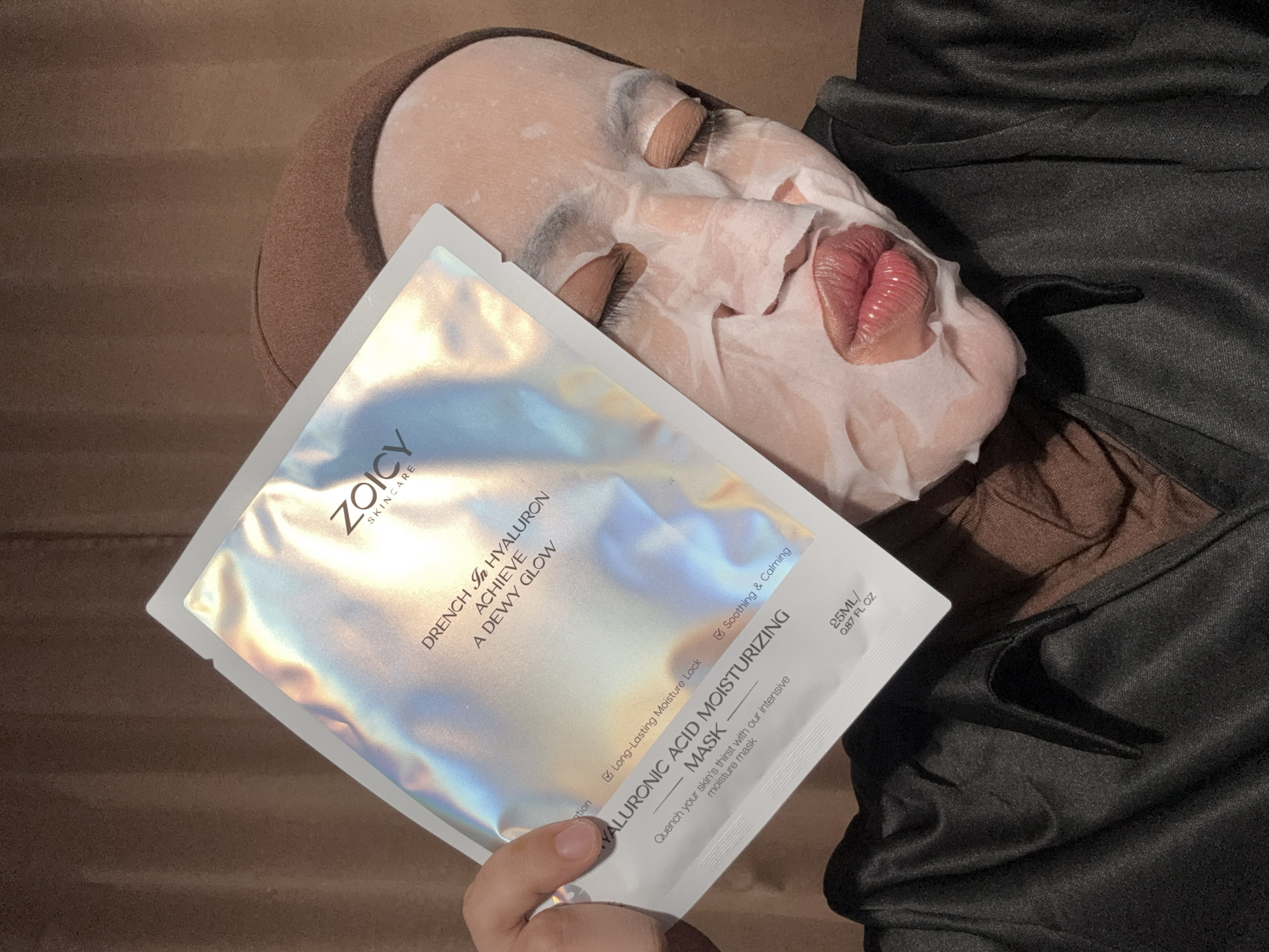 A close-up shot captures a person in Gadingrejo, Indonesia, engaging in a relaxing skincare routine, likely indoors during the evening or night due to the warm, soft lighting. The individual is lying down, with their eyes closed, indicating a state of rest and self-care. A white sheet mask is applied to their face, conforming to the contours and covering most of their features, with cut-outs for the eyes and mouth. The lips are visible, appearing hydrated.
The person is wearing a brown head covering, possibly a hijab or turban, and dark-colored clothing, which could be a robe or comfortable attire. In their left hand, they hold a pristine, holographic sheet mask packet. The packet's surface shimmers with iridescent reflections of silver, blue, purple, and green.
Visible text on the mask packet, from the brand "ZOICY SKINCARE," highlights the product's benefits: "DRENCH In HYALURON ACHIEVE A DEWY GLOW." Further details describe it as a "HYALURONIC ACID MOISTURIZING MASK" that provides "Long-Lasting Moisture Lock" and is "Soothing & Calming." The product promises to "Quench your skin's thirst with our intensive moisture mask" and contains "25ML / 0.87 FL OZ." The background is softly blurred, showing indistinct brown and black textures, suggesting a cozy environment like a bed or sofa.
