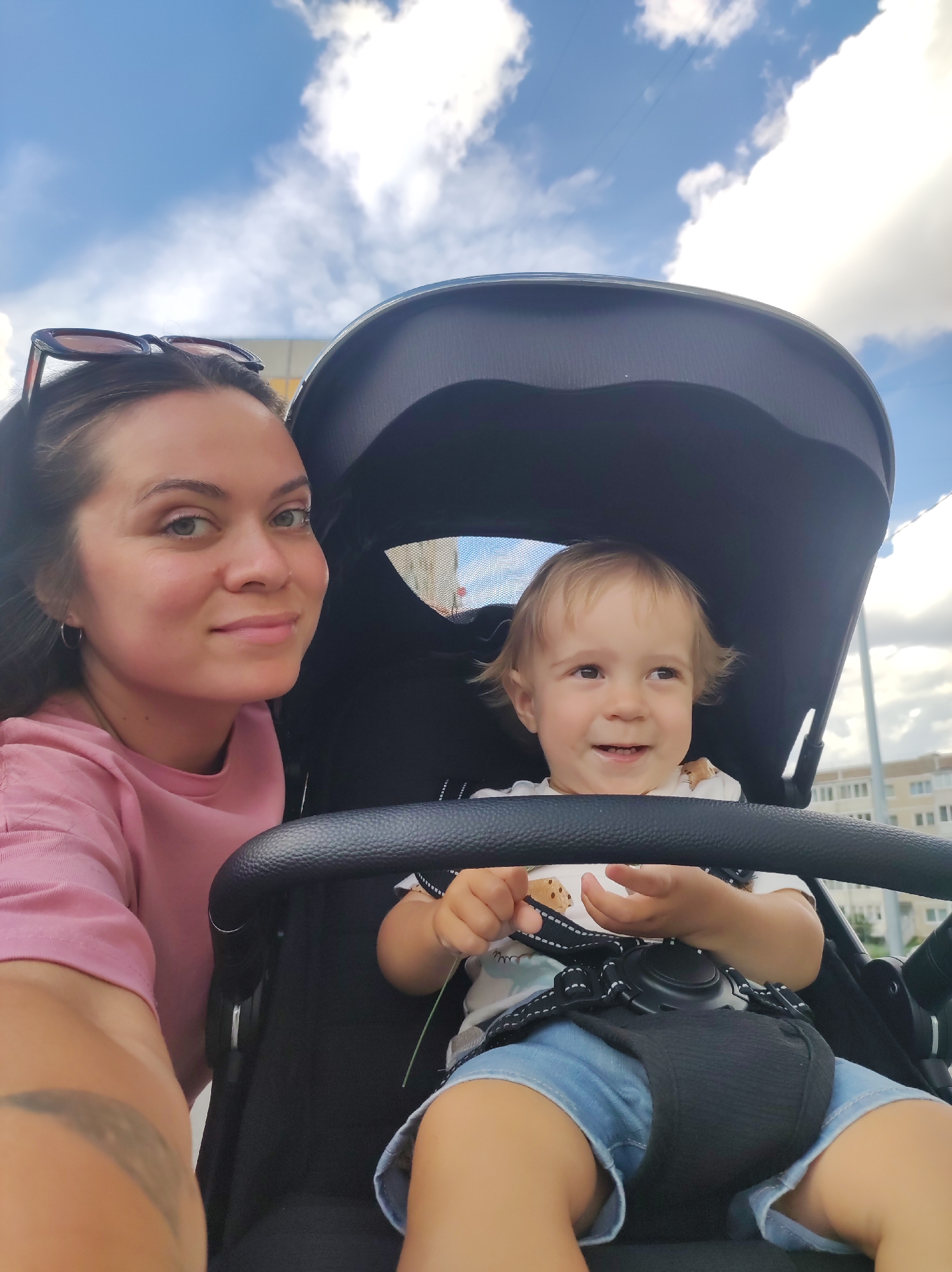 A woman and a young child are seated in a stroller. The woman is in the foreground, positioned to the left of the frame. She has dark hair pulled back and is wearing a pink t-shirt and sunglasses perched on her head. She is looking directly at the camera with a gentle smile. A tattoo is visible on her left arm.
The child is in the stroller, to the right of the woman, and is facing slightly away from the camera, looking to the right. The child has blonde hair and is wearing a white t-shirt with a graphic print and blue denim shorts. The child is smiling and holding a thin, green stem, possibly a blade of grass. The stroller has a black canopy and a black safety harness with white stitching securing the child. A black stroller bar is in the center of the frame, in front of the child.
The background shows a cloudy blue sky with fluffy white clouds. In the distance, there are buildings that appear to be apartments, suggesting an urban or suburban setting. The lighting suggests it is daytime, likely late morning or afternoon. The overall mood is cheerful and casual, indicating a pleasant outing. The location context is Chaikovsky, Russia. There is no visible text in the image.