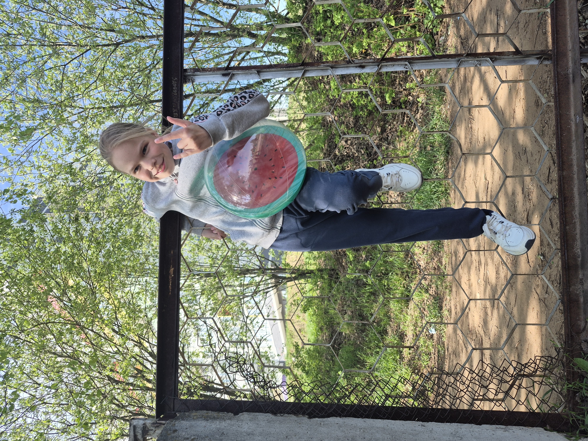 A young girl stands by a fence with a watermelon-themed frisbee, extending her hand in a "peace" sign. The fence is made of hexagonal wire mesh within a metal frame. Beyond the fence, there is a grassy area with trees and a building in the background. Through the trees, there is a partial view of a building with a white exterior. The girl is wearing a gray sweatshirt with animal print sleeves, dark pants, and white sneakers. Trees with green leaves are visible in the background behind her.