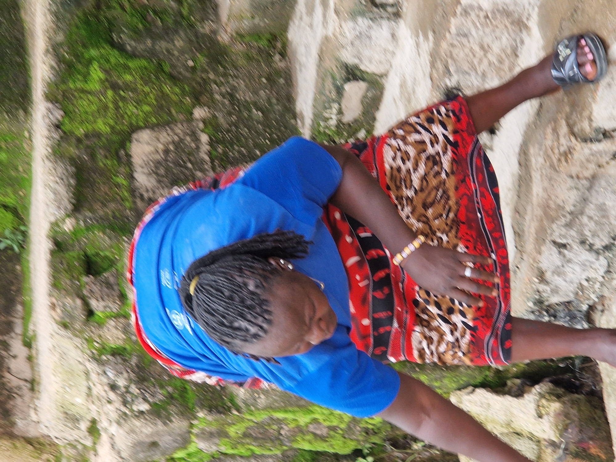 An individual is positioned on a stone staircase, leaning forward with their upper body obscured by a blue shirt. They are wearing a skirt with a red, black, and tan animal print pattern. A bracelet adorns their left wrist, and a ring is visible on their left ring finger. Their hair is styled in braids pulled back into a ponytail. The staircase and surrounding environment are covered in moss and rocks, suggesting an outdoor setting, possibly within the city of Kabong, Nigeria, given the context. The lighting indicates it is daytime.