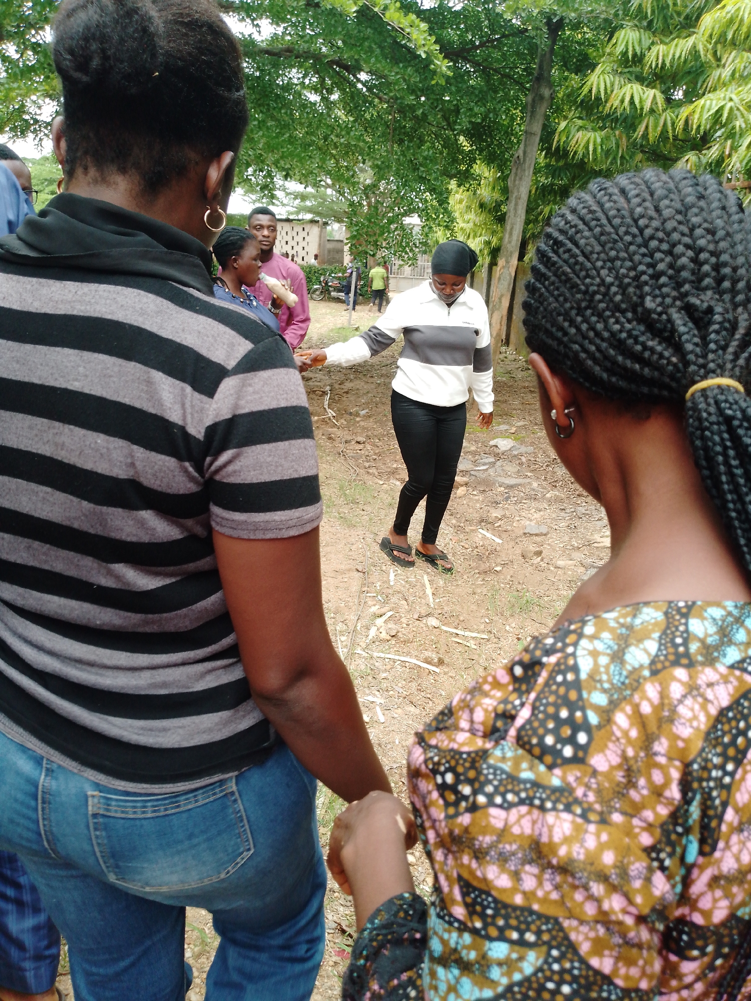 In Akwanga, Nigeria, a medium-shot image depicts a group of people in an outdoor, possibly rural, setting. The foreground is dominated by the backs of two women; the woman on the left wears a black and gray horizontally striped shirt and blue jeans, while the woman on the right is seen from the back of her head, adorned with braided hair and a vibrant, multi-colored patterned top.
In the mid-ground, a woman wearing a black headscarf, a white and gray horizontally striped long-sleeved shirt, and black pants is facing away from the camera. She is partially obscured by a person in a pink shirt standing behind her, who is holding a bottle. Another individual, possibly male, is visible in the background near a light-colored wall with ventilation blocks. Further back, a motorcycle is parked, and more people are seen in the distance, one wearing a bright green shirt.
The environment appears to be a dry, earthy area with sparse vegetation and trees providing overhead shade. The ground is covered with dirt, small stones, and some dry leaves or twigs. The lighting suggests an overcast day or shaded conditions. The composition focuses on the interaction and presence of the people within this environment, with the foreground subjects seemingly observing or interacting with those in the mid-ground.