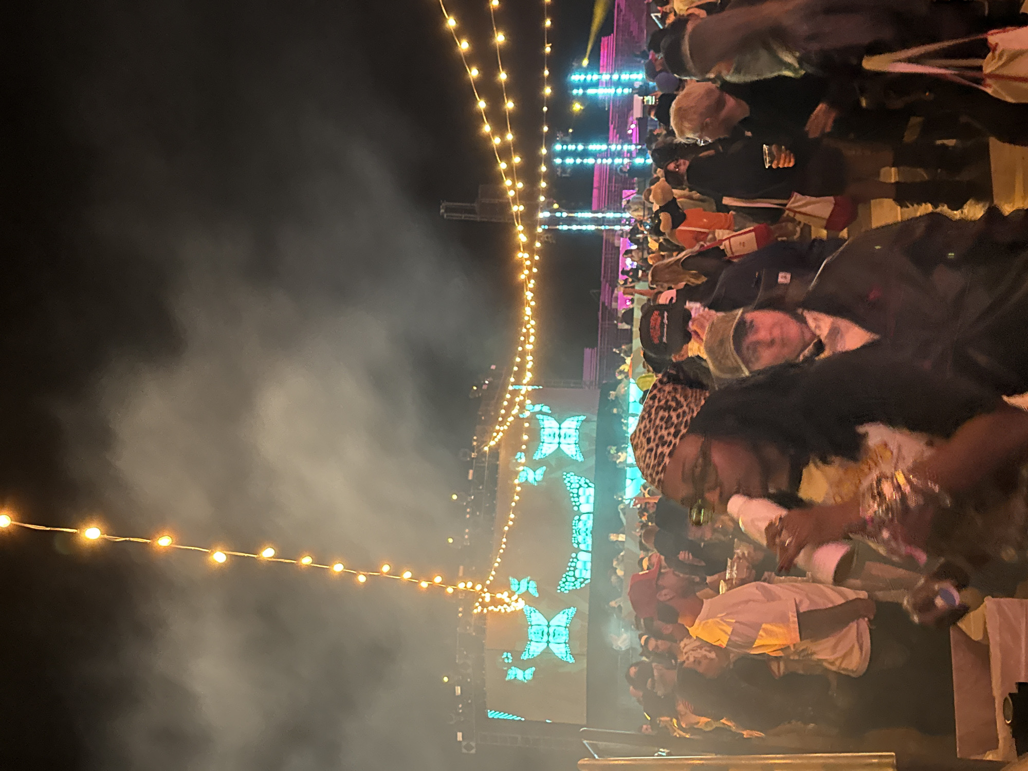 The image shows a nighttime view of what appears to be a music festival or concert in Thermal, CA, USA. A large crowd of people is visible, illuminated by a stage with bright blue butterfly-shaped light displays. A string of lights hangs overhead, stretching into the distance. Smoke or fog is present in the air. People in the foreground are identifiable, with some wearing hats or head coverings and holding cups or drinks.