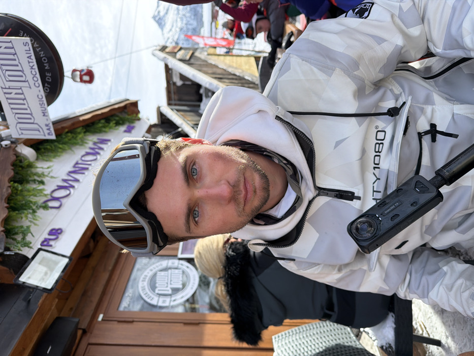 The image displays a Caucasian male subject seated outdoors in a snowy environment in front of the "Downtown" establishment, located in Les Allues, France. The male, with blue eyes and light facial stubble, wears a white and grey geometric camouflage-patterned anorak-style ski jacket with "MIGHTY1080®" branding on the chest. Grey mirrored ski goggles are positioned on his forehead. He holds a black Insta360 X4 camera on a black extension pole in his right hand, oriented towards his left side, and directly faces the primary camera. To the subject's left, partially obscured, is another individual with blonde hair, wearing a dark jacket with a black fur-lined hood, seated at an outdoor table. The background features a wooden building with a sign displaying "PUB DOWNTOWN," "BAR - MUSIC - COCKTAILS," and "SPOT DE MONTAGNE," embellished with artificial green foliage. Snowy wooden steps are visible to the right of the building. In the far background, a red gondola cabin is suspended, and snow-capped mountains rise under a clear blue sky. Snow is present on the outdoor seating and ground surfaces.