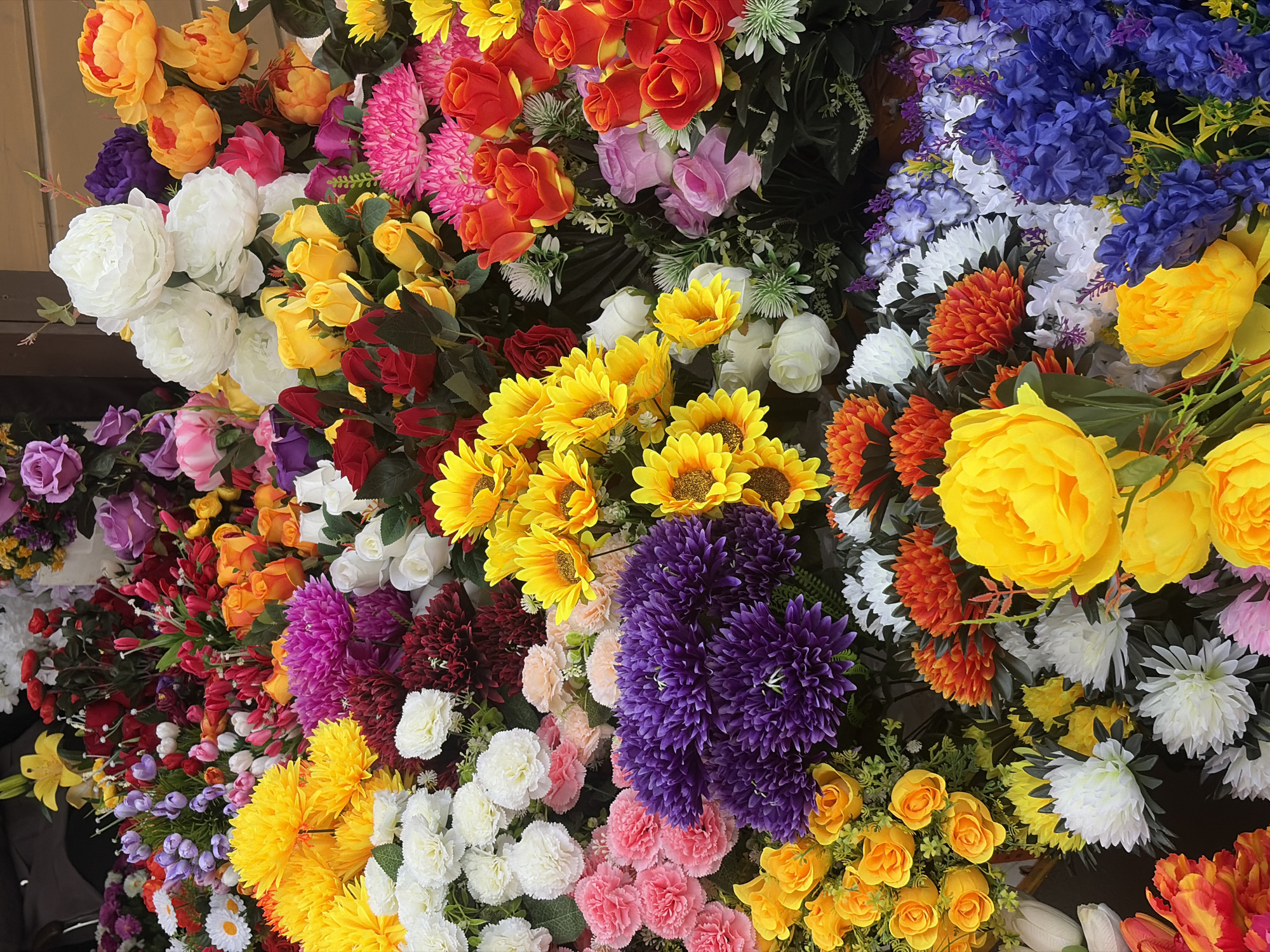 The image shows a dense arrangement of artificial flowers of various colors and types. The flowers include roses, sunflowers, hydrangeas, and peonies, with a palette ranging from orange, yellow, red, pink, purple, blue, and white. No people are visible in the frame. It is likely an indoor setting, perhaps a florist shop or market display. This image shows no indications of Saint Petersburg, Russia.