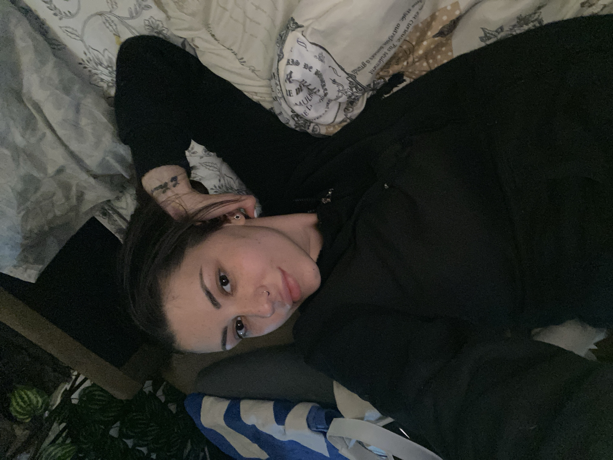 A young person is lying on a bed in a dimly lit indoor setting, gazing directly at the camera. The individual, who appears to be female, has dark hair, strong eyebrows, and a visible nose piercing. They are dressed in a dark, long-sleeved top or hoodie, and a small, illegible tattoo is noticeable on their left wrist or forearm.
The bed features patterned bedding, with one pillow displaying various textual elements. Partially discernible French phrases such as "est curieuse. Pas seulement" and "à grands" are visible on the fabric, along with "me style, autrefowances a". Other less coherent text segments like "EL E" and "[ ΦΕΥ" are also present. In the bottom left corner, some green leafy plants and a wooden surface provide a glimpse of the room's decor. A blue and white striped fabric item is also partially visible on the bed near the person.
The low lighting conditions suggest that the image was taken during the evening or night. The person's expression is neutral and relaxed, indicating a casual moment, possibly a selfie or a close-up portrait. The scene depicts a personal space, likely a bedroom, situated in Belopoptsi, Bulgaria.