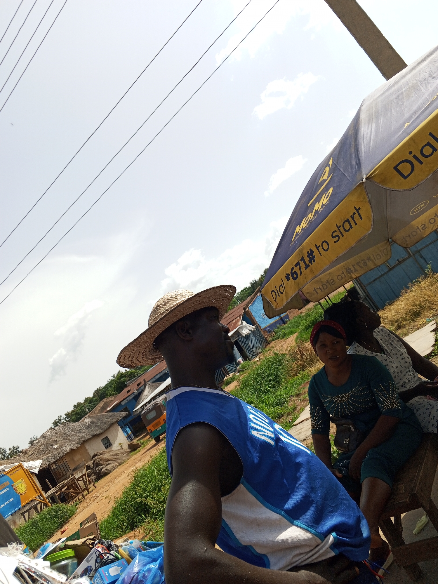 In Bukuru, Nigeria, an outdoor scene unfolds with a man wearing a straw hat and a blue sleeveless shirt in the foreground, positioned on the left, facing right. Behind him, two women are seated on a wooden bench under a large umbrella. The woman on the left wears a green top and a red headband, while the woman on the right is in a patterned white and gray outfit. The umbrella, partially visible, is blue and yellow with text indicating "Dial *671# to start" and "MOMO". The background reveals a street scene with thatched-roof buildings, green foliage, and a sandy dirt road. A yellow auto-rickshaw is visible further down the road. In the lower foreground, there are various items, including plastic bags containing goods, a jar, and other assorted objects. Overhead, power lines crisscross against a bright, cloudy sky.