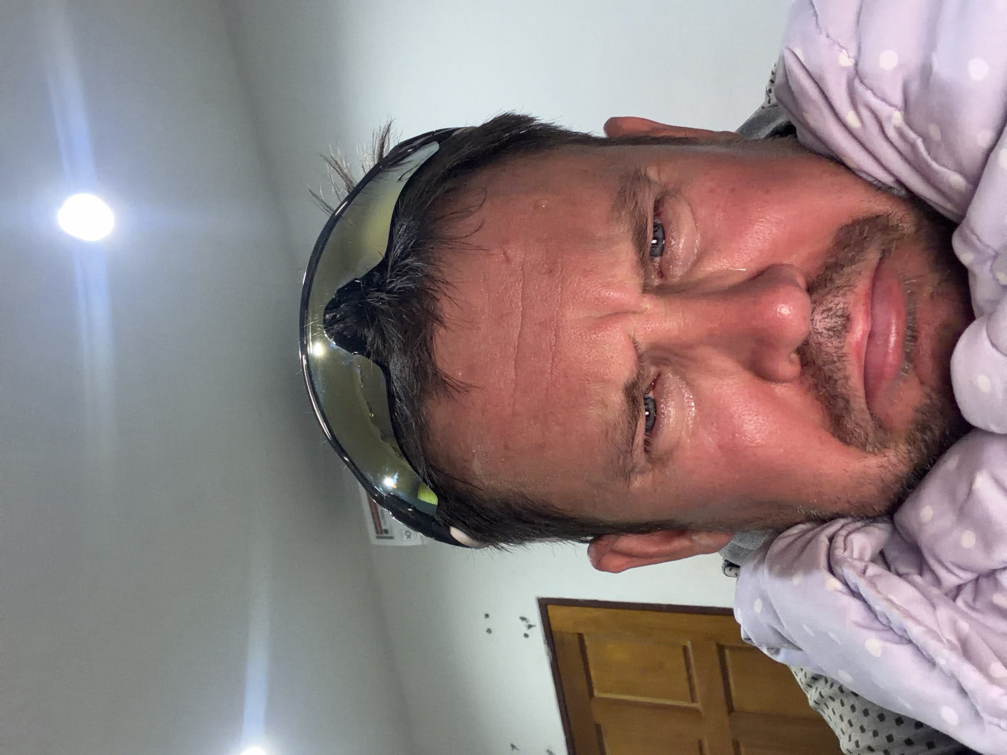 The image shows a close-up, head-and-shoulders shot of a fair-skinned man, appearing to be in his late 30s or early 40s, with dark hair and stubble, looking directly at the camera. He has sweat on his forehead and is crying, with tears visible on his cheeks and around his eyes. He is wearing large, dark-tinted sports sunglasses pushed up on his forehead. He is wrapped in a light purple blanket with white polka dots. The background consists of a plain, light-colored wall with some faint marks and two overhead light fixtures. To the left of the man, a portion of a wooden door is visible. The scene appears to be indoors, possibly in a residential setting. The man's expression suggests distress or sadness. No specific landmarks identifiable as being in Baan Mae Nam, Thailand are present in the image.