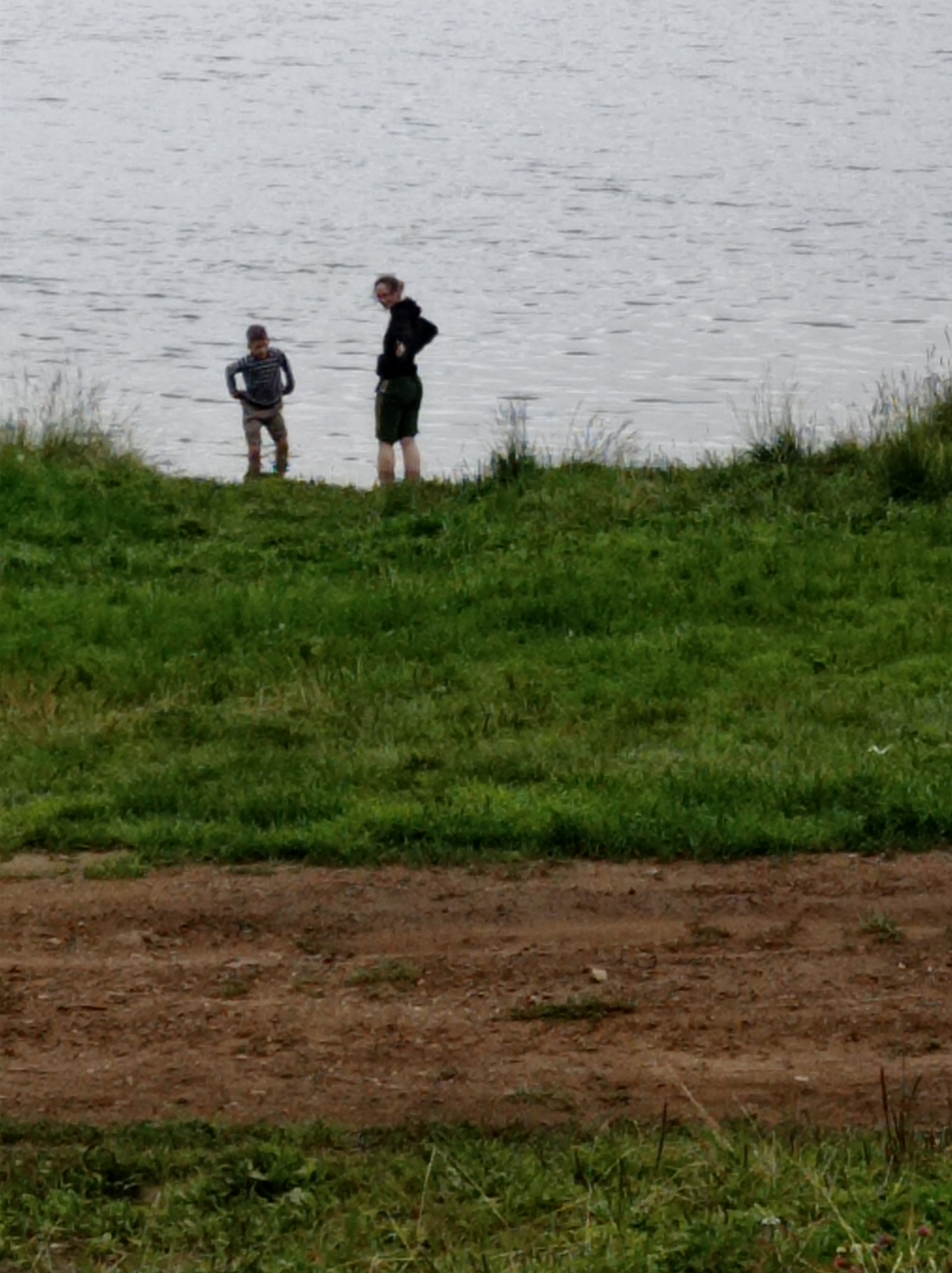 In the city of Degtyarsk, Russia, two individuals are observed near a body of water. The scene depicts a grassy embankment leading down to the water's edge. On the embankment, a young boy, wearing a striped shirt and shorts, stands facing towards the water. To his right, a person, likely an adult, is positioned with their back mostly to the camera, wearing a dark hooded jacket and shorts. The water appears calm with ripples. The ground in the foreground is a mix of dirt and grass.