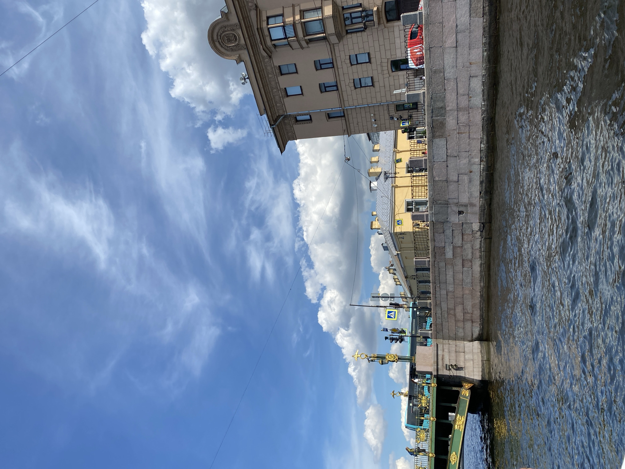 The image shows a view of a canal in Saint Petersburg, Russia, with a bridge and buildings on the embankment under a partly cloudy blue sky. The canal water is dark and rippled, reflecting the sky and buildings. On the left, a green bridge with ornate gold detailing spans the canal. A blue bus is visible on the bridge, and pedestrians are walking on the sidewalk. Traffic lights and pedestrian crossing signs are present on the embankment.
Dominating the right side of the image is a light brown, multi-story building with classical architectural details, including a decorative cornice and arched balconies. Adjacent to it is a lower, yellow building with a grey roof and yellow decorative fencing. The embankment is constructed of large, grey stone blocks. The sky features scattered white cumulus clouds against a bright blue backdrop, with thin wispy clouds higher up.
