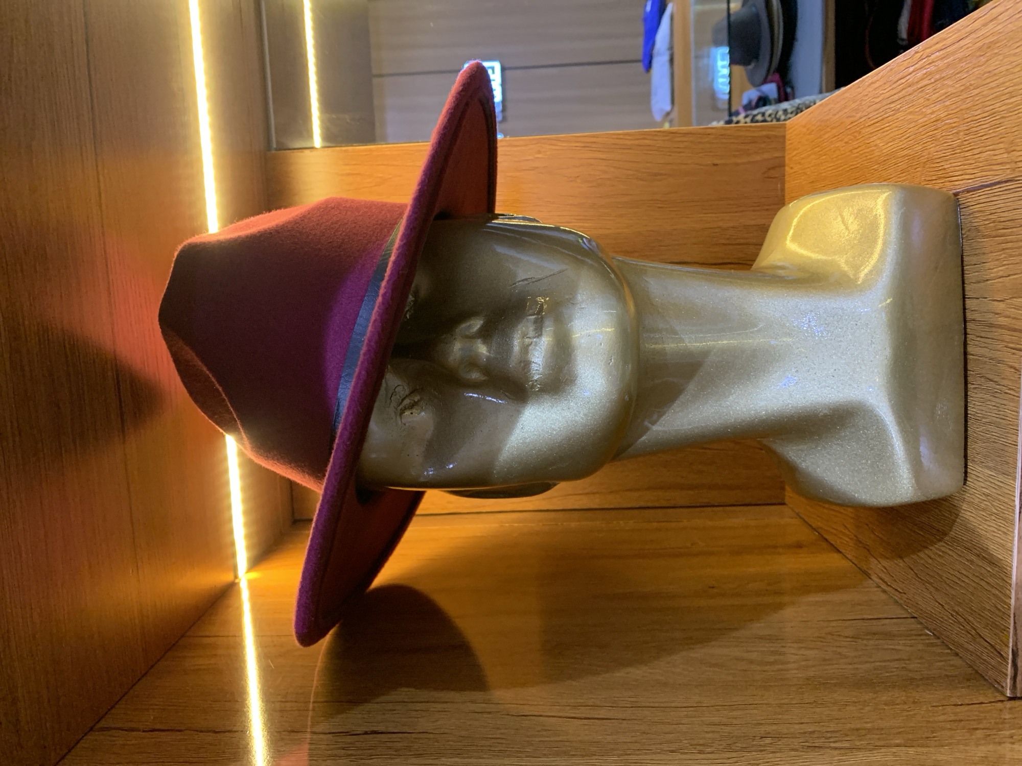 A golden mannequin head wearing a burgundy fedora hat with a dark band is positioned in the center of the frame. The mannequin is set against a backdrop of wooden shelving, illuminated by strip lighting. The overall scene suggests a retail display, possibly in a hat shop. The specific location in Gura Top, Nigeria, is not evident from the visual information provided.