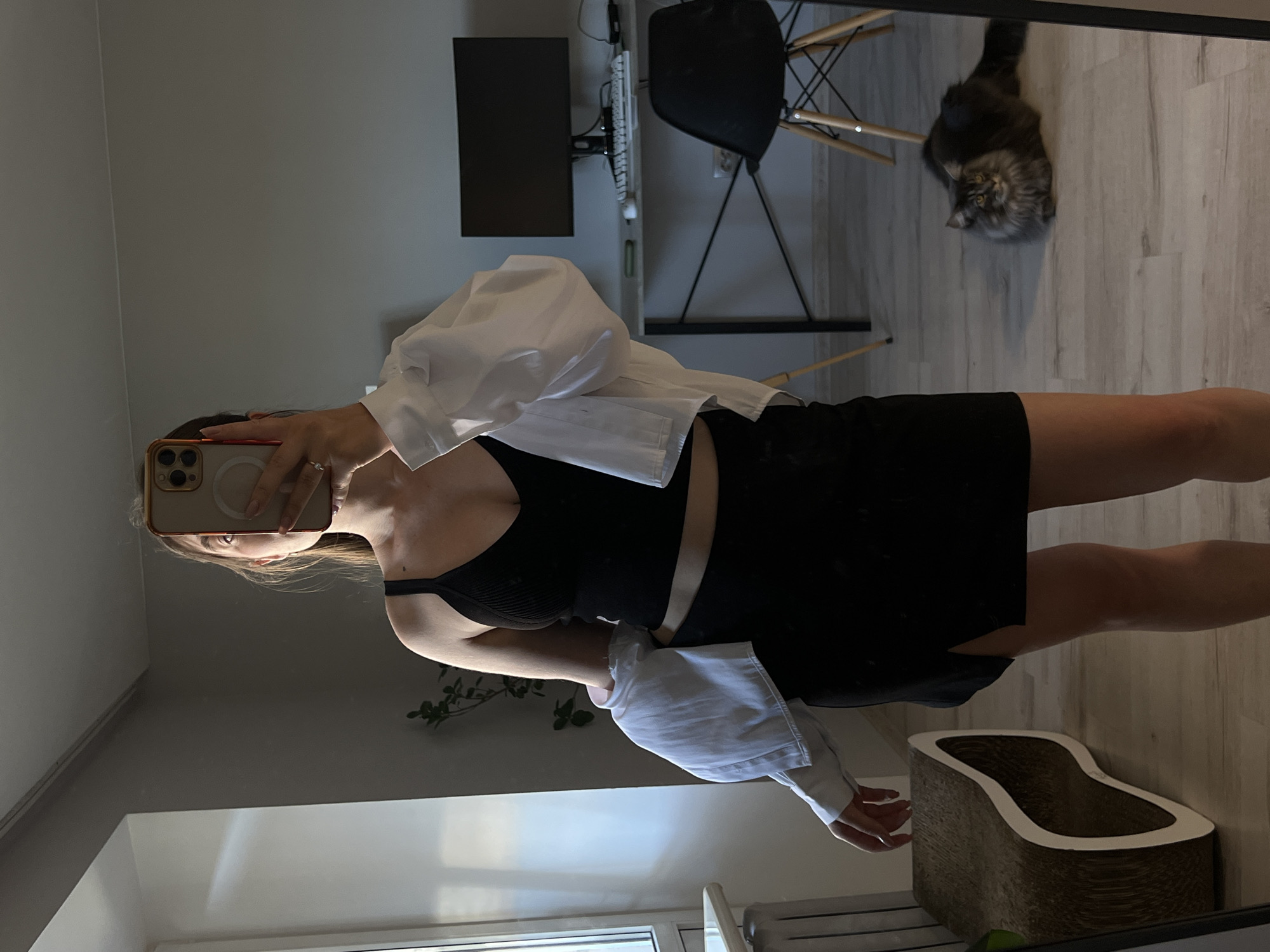 A young woman is taking a mirror selfie in what appears to be a bedroom or living space. She is wearing a black crop top and a black skirt with a slit on the side. A white shirt is draped over her shoulders. She is holding a phone in her right hand, obscuring her face. A cat is lying on the floor in the background, to the right of the woman. The room has light-colored walls and a wooden floor. There is a desk with a computer monitor and keyboard, and a black chair behind it. A window is visible on the left side of the image. The overall impression is of a casual, domestic setting. The presence of a cat and the overall styling suggest a modern, possibly urban environment. The specific location within Ternopil, Ukraine cannot be determined from the visual information alone.
