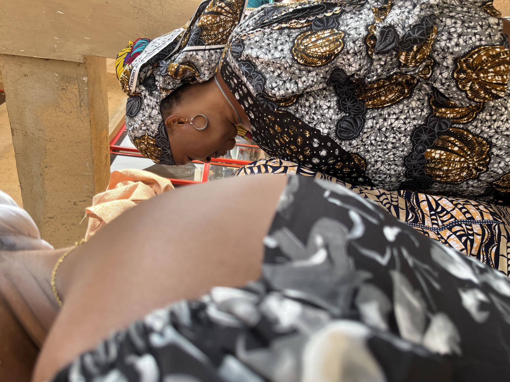 In Bukuru, Nigeria, a woman is captured in profile, wearing a traditional patterned headwrap and an equally ornate garment. The fabric of her clothing and headwrap features a complex design of black, white, and gold motifs, including what appears to be representations of shells or seed pods. She wears a silver hoop earring in her left ear and a delicate silver chain necklace. Her expression is neutral as she looks downwards. The background suggests an outdoor setting, possibly a market or an open-air gathering, with a beige, textured wall and a hint of a red structure in the distance. The presence of another person, partially visible and out of focus in the foreground, indicates a social context. The lighting suggests it is daytime.