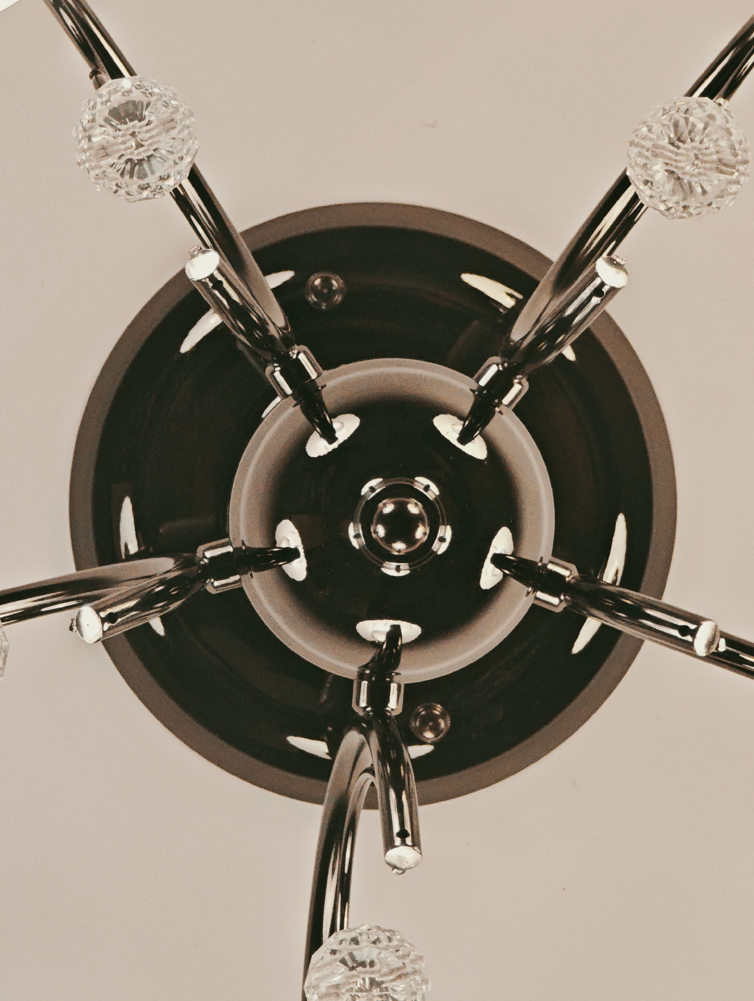 This is a close-up, overhead shot of a modern chandelier. The chandelier has a dark, circular base from which several polished metal arms extend outwards. Each arm is tipped with a clear, faceted crystal orb, catching the light. The central part of the chandelier features a layered design with a polished metal fixture. The background is a plain, light-colored surface, likely a ceiling. The lighting appears to be ambient, casting soft reflections on the metallic surfaces. The image is a still shot, with no activity or event taking place. The focus is entirely on the intricate details of the chandelier's design. There is no visible text in the image.