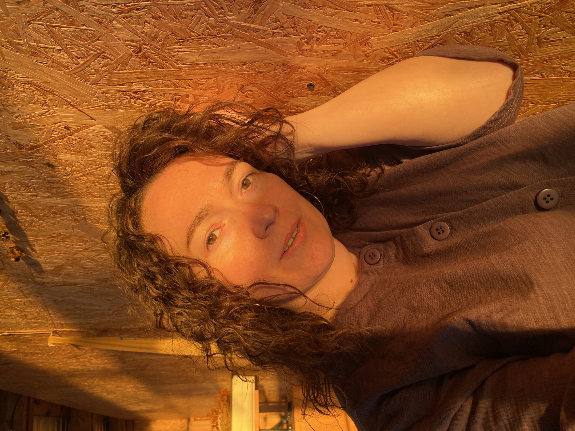 A woman with curly brown hair and fair skin is lying down in what appears to be an interior space with plywood walls. She is wearing a brown button-up shirt. Her arm is raised, and her hand is near her head. Light, possibly sunlight, is illuminating her face and the surrounding area. The location, Ольховка, Russia, is not discernible from the image.