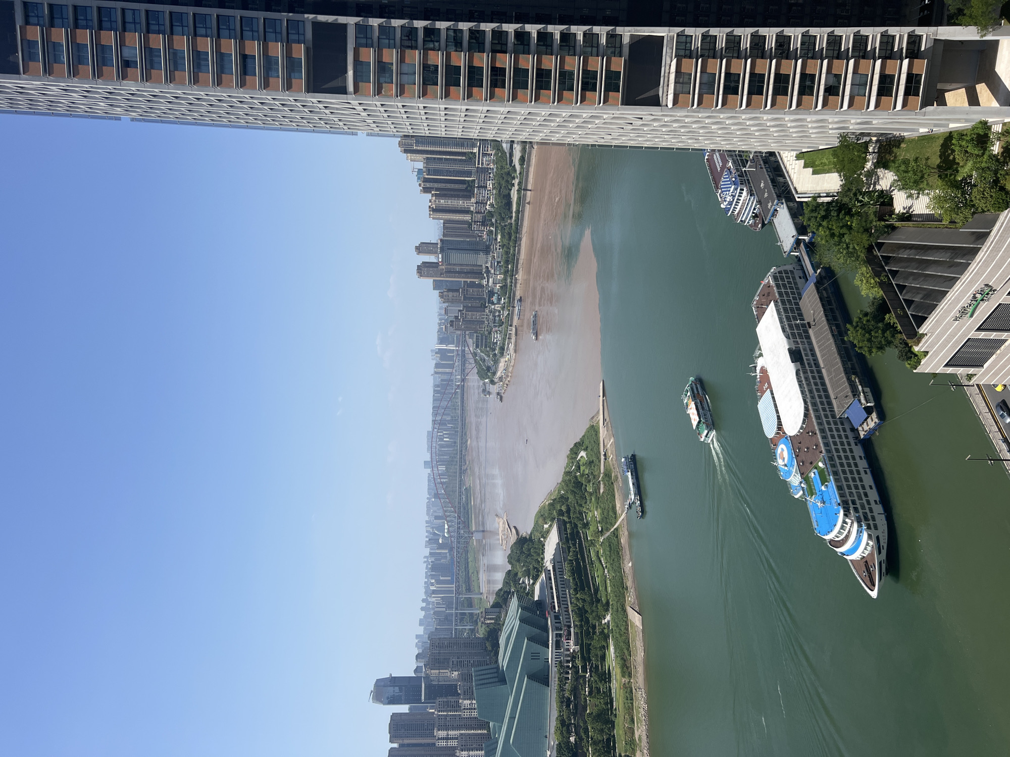 This is a high-angle, panoramic photograph of the Yuzhong District in Chongqing, China, on a clear, sunny day. The foreground is dominated by the right side of a modern, tall skyscraper with a beige and glass facade. The middle ground features the confluence of two rivers, one with green, clear water and the other with muddy, brown water, creating a distinct color demarcation. Several large passenger boats, appearing to be cruise ships, are docked or moving on the green river. One large white cruise ship with blue accents is prominently positioned in the lower left quadrant of the frame. In the distance, a sprawling cityscape with numerous high-rise buildings stretches across the horizon. A large bridge with red arches spans the river in the far distance, connecting different parts of the city. The sky is a clear, vibrant blue. The overall scene depicts a bustling urban environment situated alongside a significant river system.