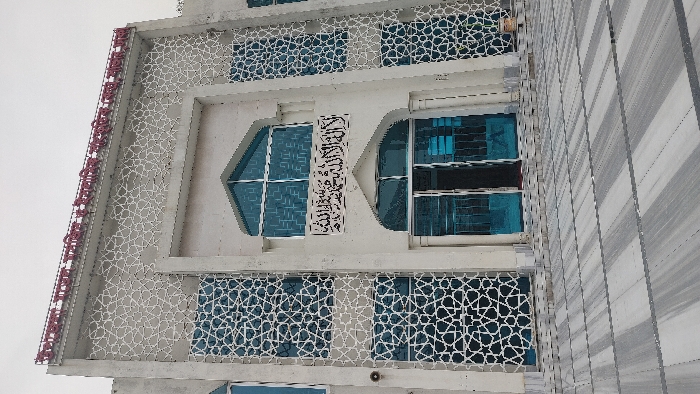 The image displays the front facade of the Upazila Social and Cultural Center, situated in Savar, Bangladesh. The building is a light-colored, multi-story structure with a modern architectural style incorporating Islamic design elements. Its exterior features extensive vertical sections of white geometric lattice patterns overlying or integrated with large, blue-tinted glass windows. A central, recessed arched entryway contains partially open double blue-tinted doors, providing a glimpse into the dark interior. Above this entrance, a horizontal panel displays black Arabic calligraphy, which translates to the Shahada ("There is no god but Allah, Muhammad is His Messenger"). A large, arched, multi-pane window is positioned above the calligraphy. Along the top edge of the building, red Bengali script identifies the structure as an "Upazila Social and Cultural Center." The foreground consists of a meticulously paved plaza composed of large, rectangular tiles with alternating light gray and white stripes, laid horizontally, leading directly to the building's entrance. The sky overhead is overcast. No individuals, vehicles, or active scenes are present within the frame.