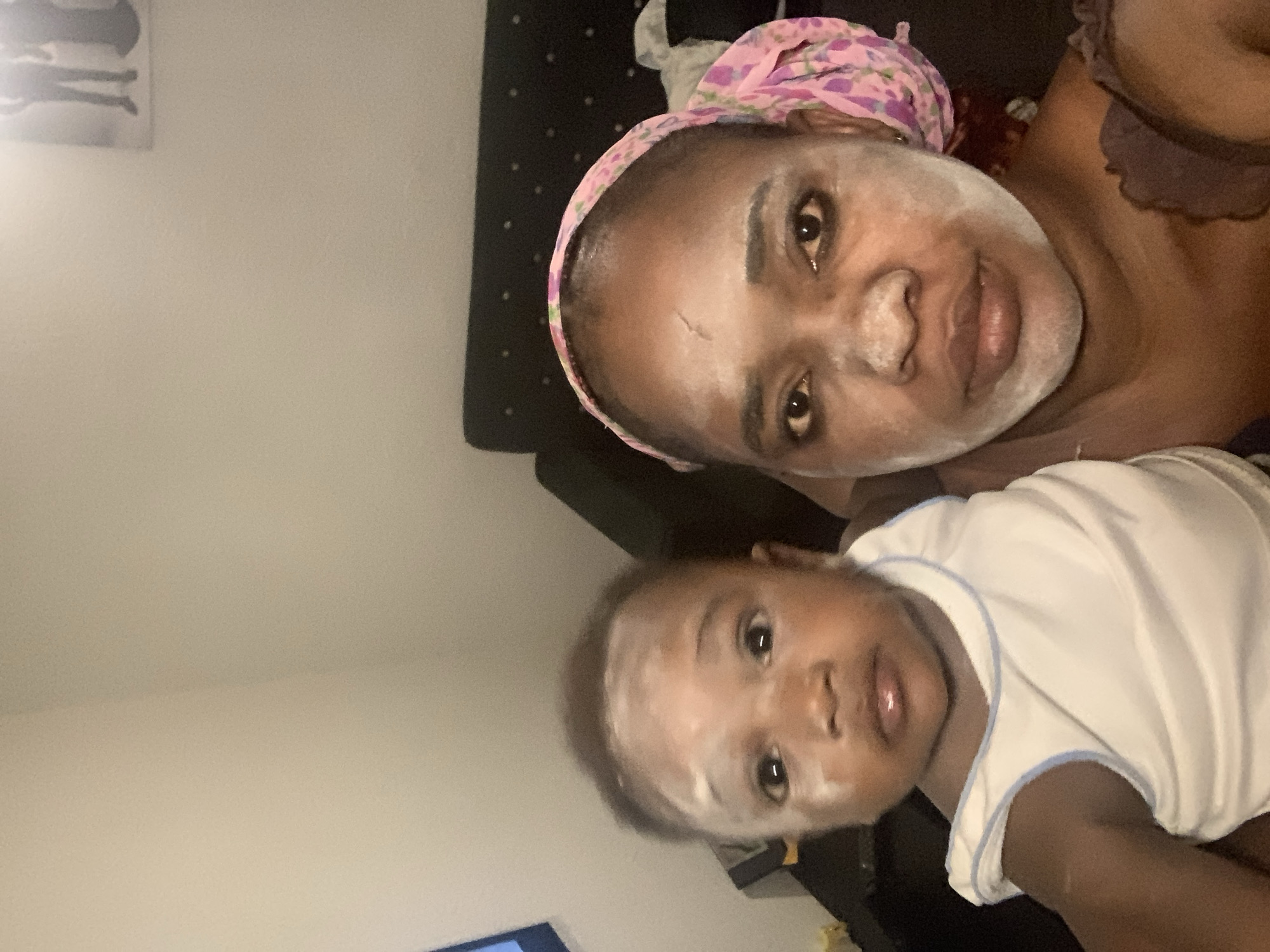 A selfie shows a woman and a baby lying on what appears to be a bed. Both have a white powdery substance on their faces. The woman wears a pink and purple headscarf. The baby is wearing a white shirt with light blue trim. The background includes a wall with an abstract picture, and a black headboard. The location is most likely a private residence. There are no visible features confirming the city of Barakin Akawo, Nigeria.