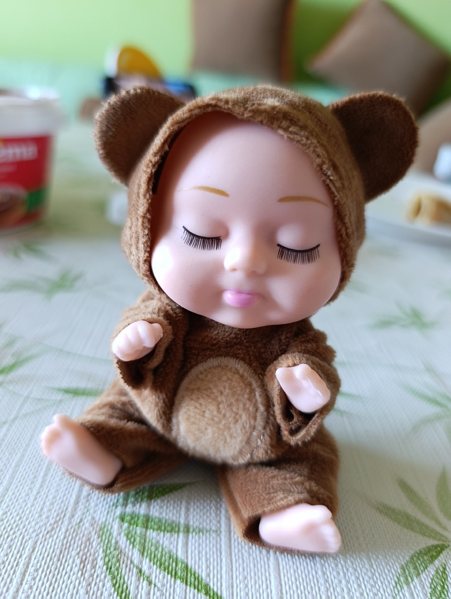 A doll wearing a brown bear suit is sitting on a table. The table is covered with a green and white patterned tablecloth. In the background, there is a tub of food and some furniture. The doll has closed eyes and a small pink mouth. The image was likely taken in Plovdiv, Bulgaria.