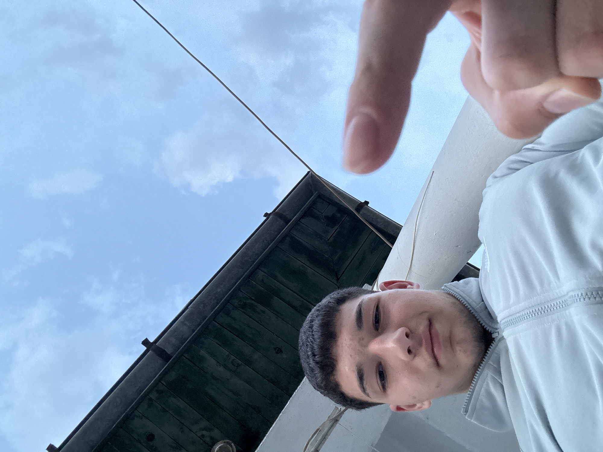 A low-angle selfie featuring a young man with dark hair, light complexion, and wearing a light gray jacket. His hand and finger are raised into the frame obscuring part of the sky. Behind him is a roof edge and a building possibly located in Zlatograd, Bulgaria based on the surrounding architecture. The sky is cloudy. A cable or wire runs diagonally across the top left of the image.