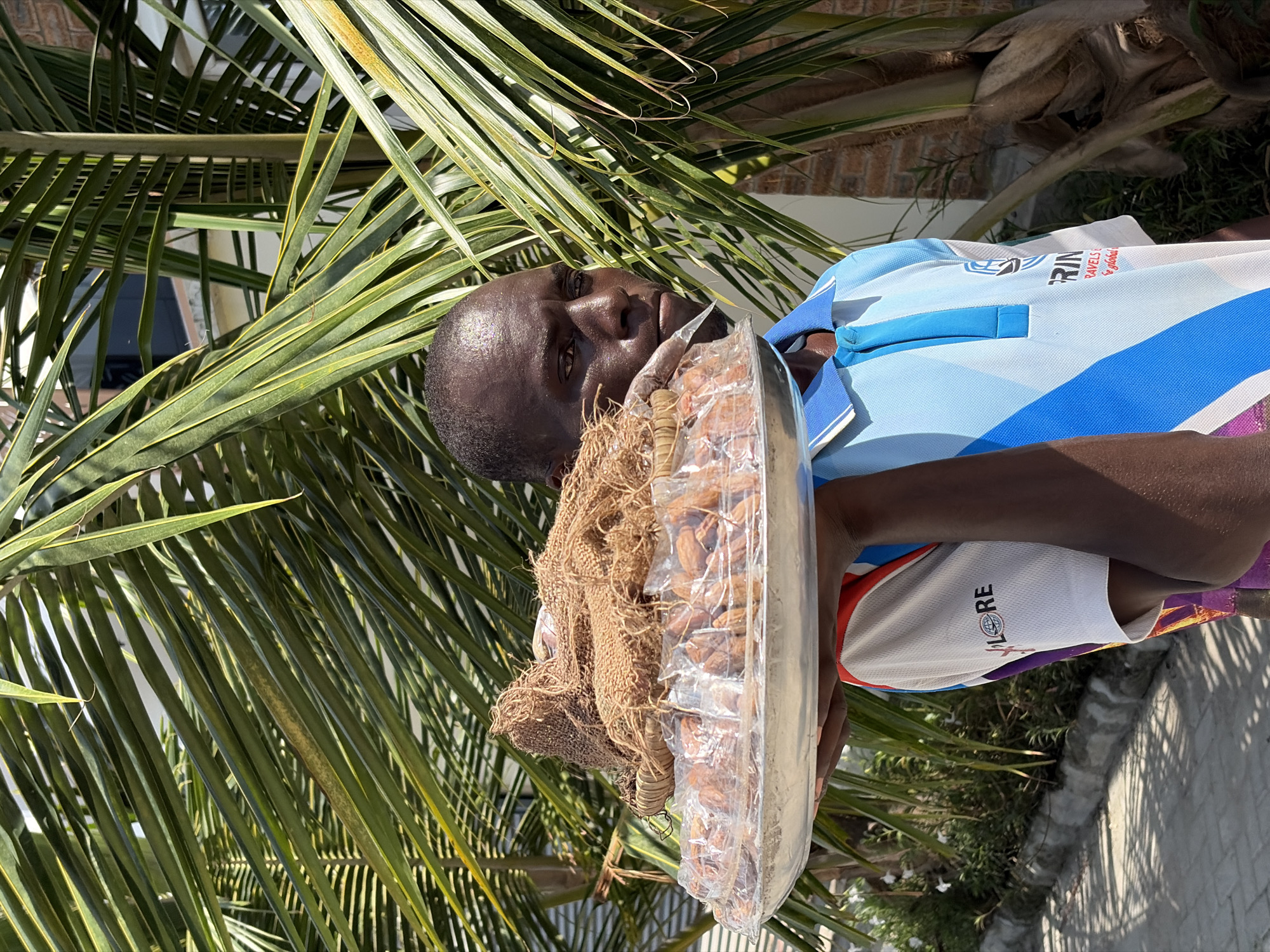A man is standing outdoors near palm fronds in Igboefon, Nigeria, holding a clear plastic container filled with brown nuts. The container is partially covered by a coarse, brown material. The man is wearing a light blue and white polo shirt. A building and a paved surface are visible in the background.
