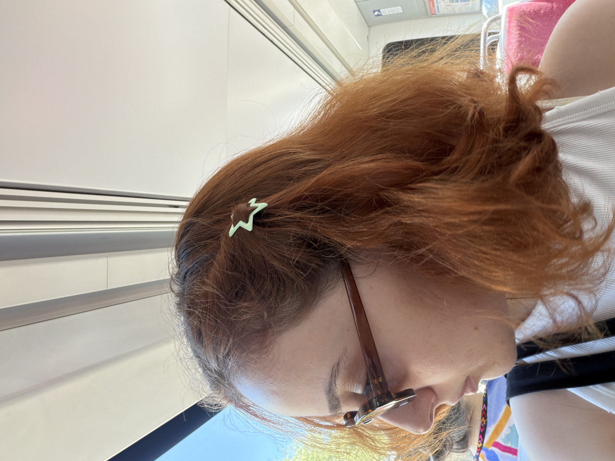 The image shows a close-up profile of a person with reddish-brown hair. A light green star-shaped hair clip is visible in their hair. The person is wearing glasses with brown frames and a white shirt with black stripes. The background appears to be an interior, possibly a train or bus, with white walls and horizontal metallic elements. Outside the window, a bright blue sky is visible, suggesting it is daytime. The person's hair is somewhat messy, with some strands out of place. The image is taken from a slightly elevated angle, looking down at the person. The overall impression is casual, possibly during travel. There is no visible text in the image.