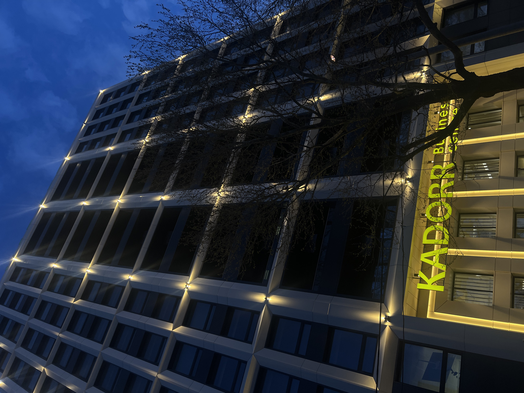 A modern, high-rise building dominates the scene under a dark blue, cloud-streaked sky at night or deep twilight. The setting is urban, specifically in Odesa, Ukraine. A large, bare-branched deciduous tree stands prominently in front of the building, its dark branches creating an intricate silhouette that partially obscures the structure.
The building's facade features a contemporary design with a grid-like pattern of recessed rectangular windows, most of which appear dark, reflecting the night. Numerous warm-toned exterior lights illuminate the building; individual spotlights cast upward beams from ledges, and glowing strip lights define some of the vertical and horizontal architectural lines.
On the right side of the building, a large, bright green illuminated sign reads "KADORR" in tall, vertical letters. Beneath "KADORR," the words "Business Centre" are visible in smaller, white vertical text, identifying the building's function. No people or specific activities are visible, emphasizing the architectural presence and the interplay of light and shadow in the urban nightscape. The overall mood is serene and highlights the building's modern aesthetic.