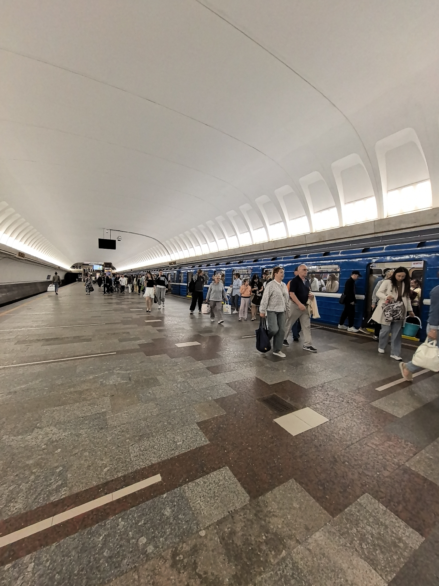 The image shows a busy subway platform in a city. The platform is wide with a tiled floor made of large, square tiles in shades of grey and brown. A long, blue subway train is stopped at the platform, with its doors open. Many people are seen disembarking and boarding the train, as well as waiting on the platform. Some individuals are carrying bags and luggage. The ceiling of the station is high and arched, with a white, curved design and rows of lights. A black screen is mounted on the ceiling in the distance. The overall scene depicts a typical urban commute during the day.