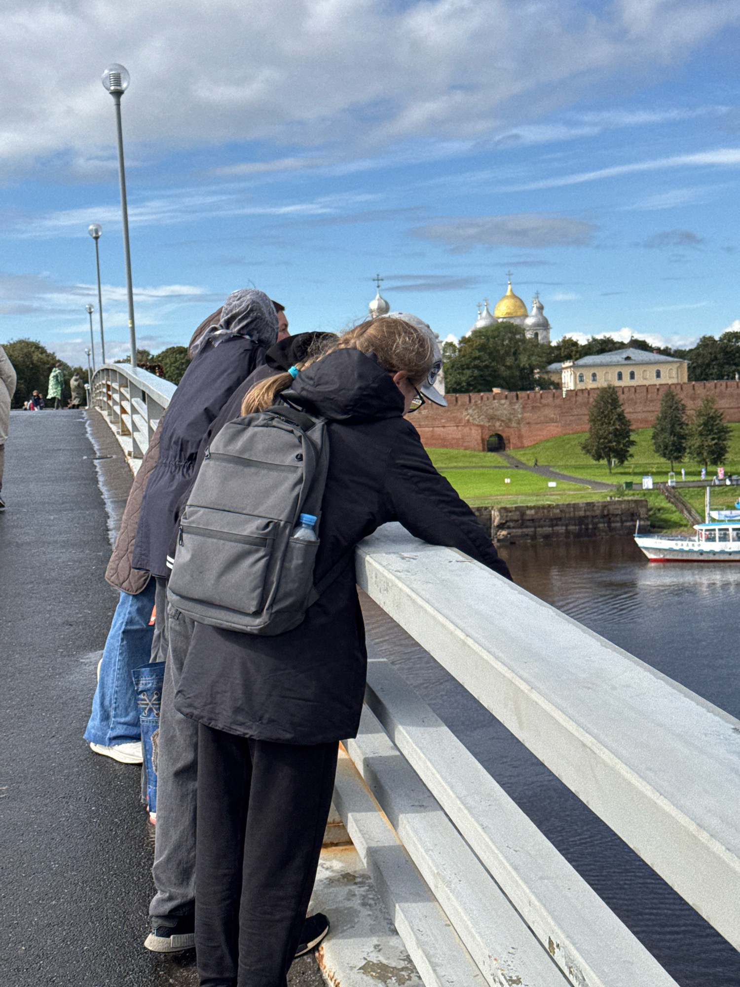 This image depicts a group of people standing on a bridge overlooking a river in Veliky Novgorod, Russia. The people are looking towards a large, historic complex with red brick walls and domed churches in the distance. In the foreground, a person wearing a black jacket and a grey backpack is leaning on the bridge railing, observing the view. Other individuals, dressed in casual attire, are standing alongside. The bridge appears to be a pedestrian walkway, with lampposts lining its side. The river reflects the cloudy sky, and a boat is visible on the water. The overall scene suggests a tourist or recreational outing, with the individuals taking in the historical and scenic beauty of the city.