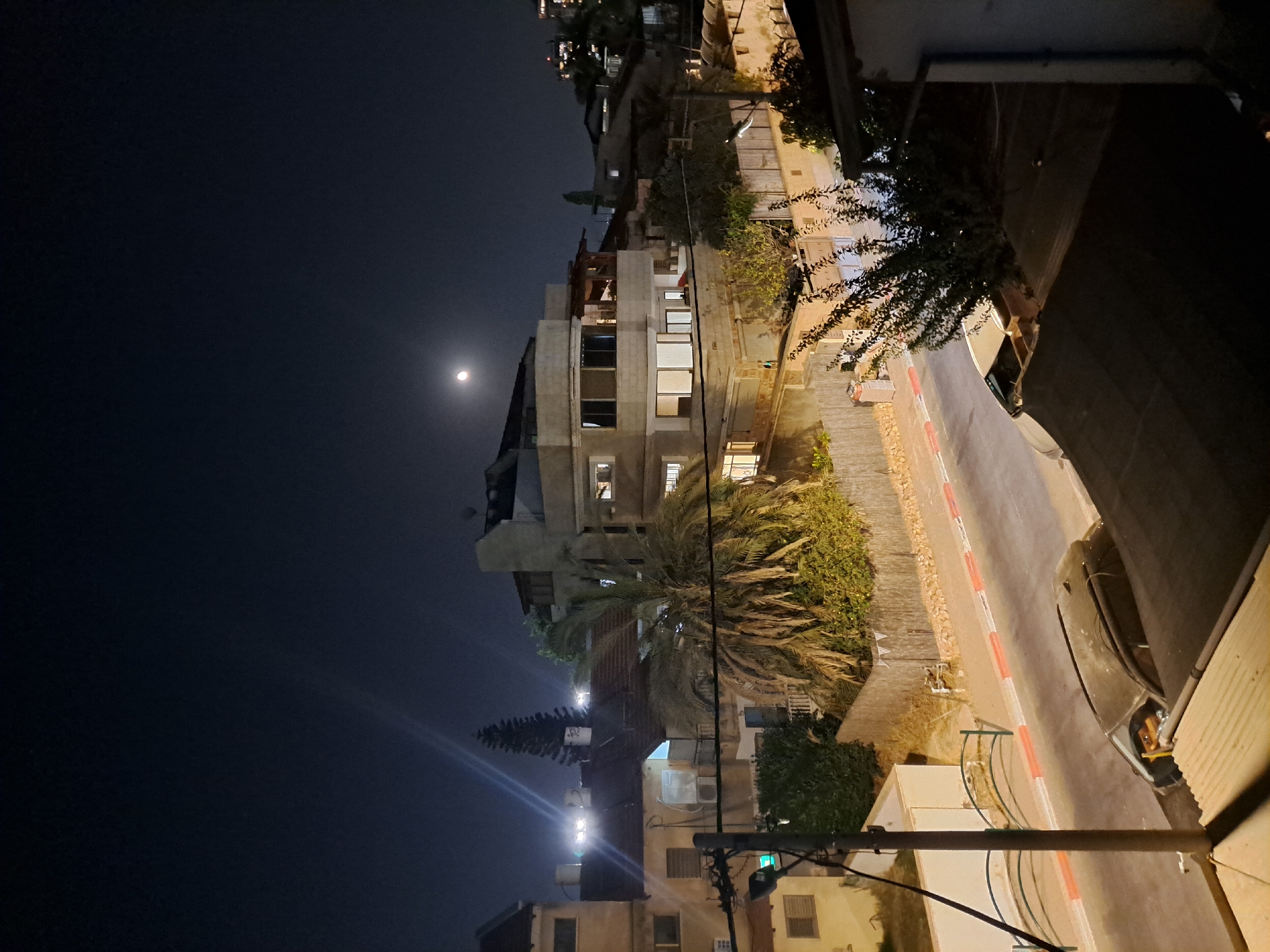 The image depicts a nighttime street scene in Ramat HaSharon, Israel, under a dark sky illuminated by a crescent moon. The foreground shows the dark roof of a building with some foliage. A street runs through the midground, with a parked dark-colored car visible on its left side. The street is bordered by a sidewalk with red and white markings. Adjacent to the sidewalk is a wooden fence, behind which are various residential buildings and lush vegetation, including a prominent palm tree and a tall evergreen. Streetlights and internal building lights cast a glow on the scene. One building in the mid-right is brightly lit from within. The overall atmosphere is quiet and residential.
