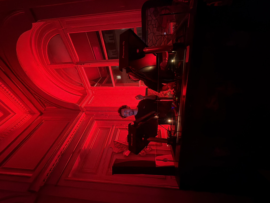 An interior room, suffused with deep red lighting, forms the setting. A male subject, possessing dark curly hair and a beard, stands centrally behind a professional DJ console. He wears a dark short-sleeved shirt, and his right hand is elevated with the index finger extended, poised over equipment. The DJ setup comprises at least two Pioneer DJ branded media players and a central mixer, all mounted on a dark, elevated counter. A black speaker unit is positioned to the left of the console. The room's walls are adorned with prominent ornate classical moldings and a decorative cornice, indicative of 19th-century Haussmannian architecture. A large arched window on the right reveals an obscured view of an adjacent building's façade under low external light, consistent with nighttime. Based on the distinctive Haussmannian architectural style, the location is identified as within the city of Paris, France, likely a historic building converted for an event or club.