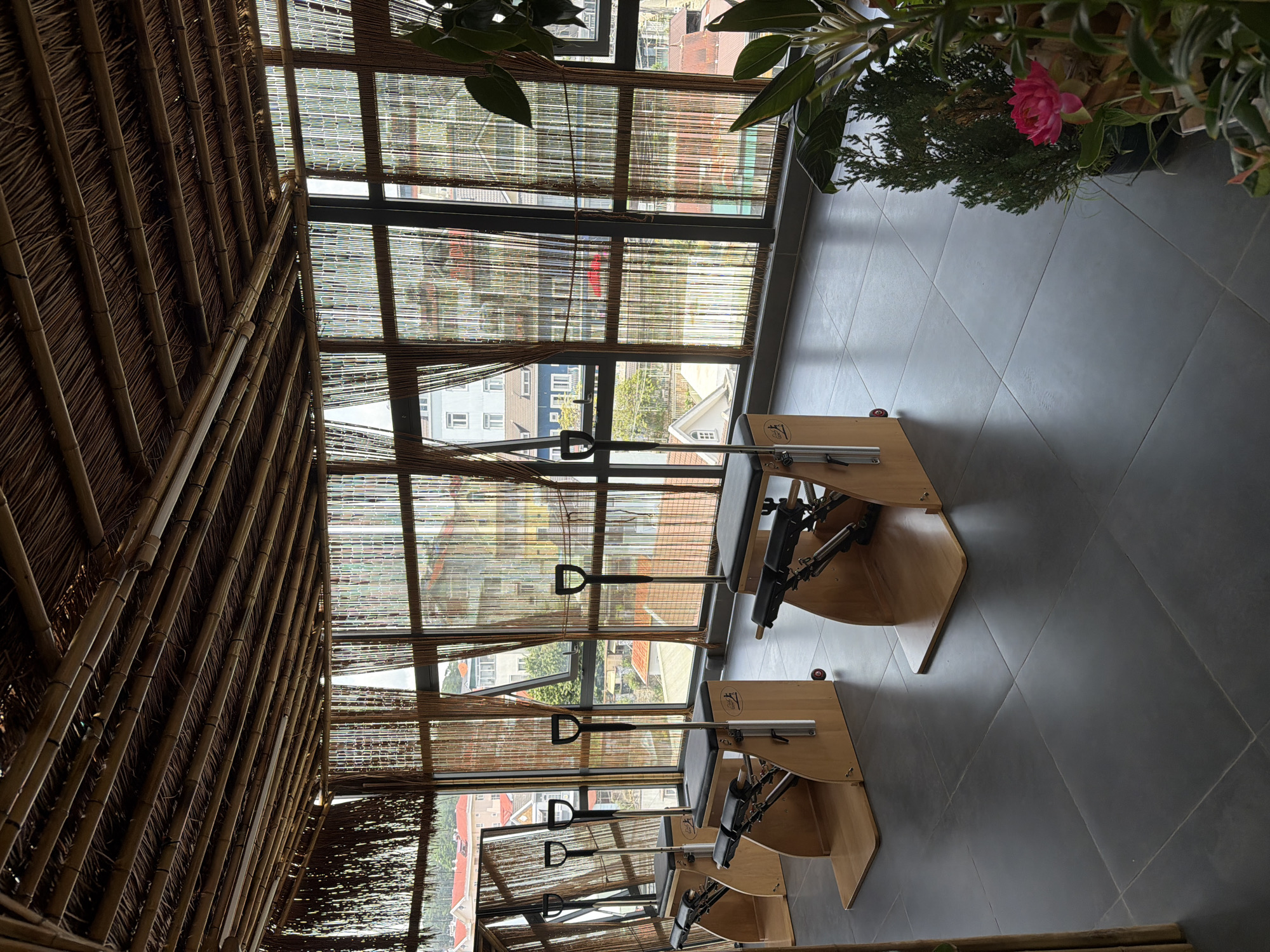 This image depicts the interior of a Pilates studio in Da Lat, Vietnam. The room is characterized by its natural materials, with a bamboo ceiling and woven bamboo blinds covering large windows that offer a view of the city's rooftops and buildings. In the foreground, three Pilates reformers are arranged neatly on a dark tiled floor. Each reformer is made of wood and metal, featuring a black padded seat and adjustable handles. To the right, several potted plants, including a pink flower, add a touch of greenery to the space. The overall atmosphere is serene and minimalist, suggesting a focus on wellness and mindful movement.