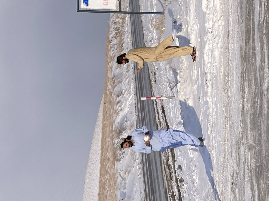 The image captures a snowy scene in Churmian, Pakistan, under an overcast sky. Two adult males stand on snow-covered ground adjacent to a road. The man on the left wears a light blue shalwar kameez and a patterned cap, holding a spherical object, consistent with a snowball, in both hands. He has dark hair and a beard, and looks towards the right. The man on the right wears a light brown shalwar kameez, dark sandals, and sunglasses, also with dark hair and a beard. He stands with his right leg slightly lifted and his right hand clenched. Between the two individuals, two red and white striped road markers are visible in the snow. A two-lane road, appearing wet and partially covered in snow, runs horizontally across the mid-ground. A partial road sign with a metallic frame is visible on the far right. In the background, hills with patches of dry, light brown vegetation are covered in snow, and overhead power lines are visible against the left hillside.