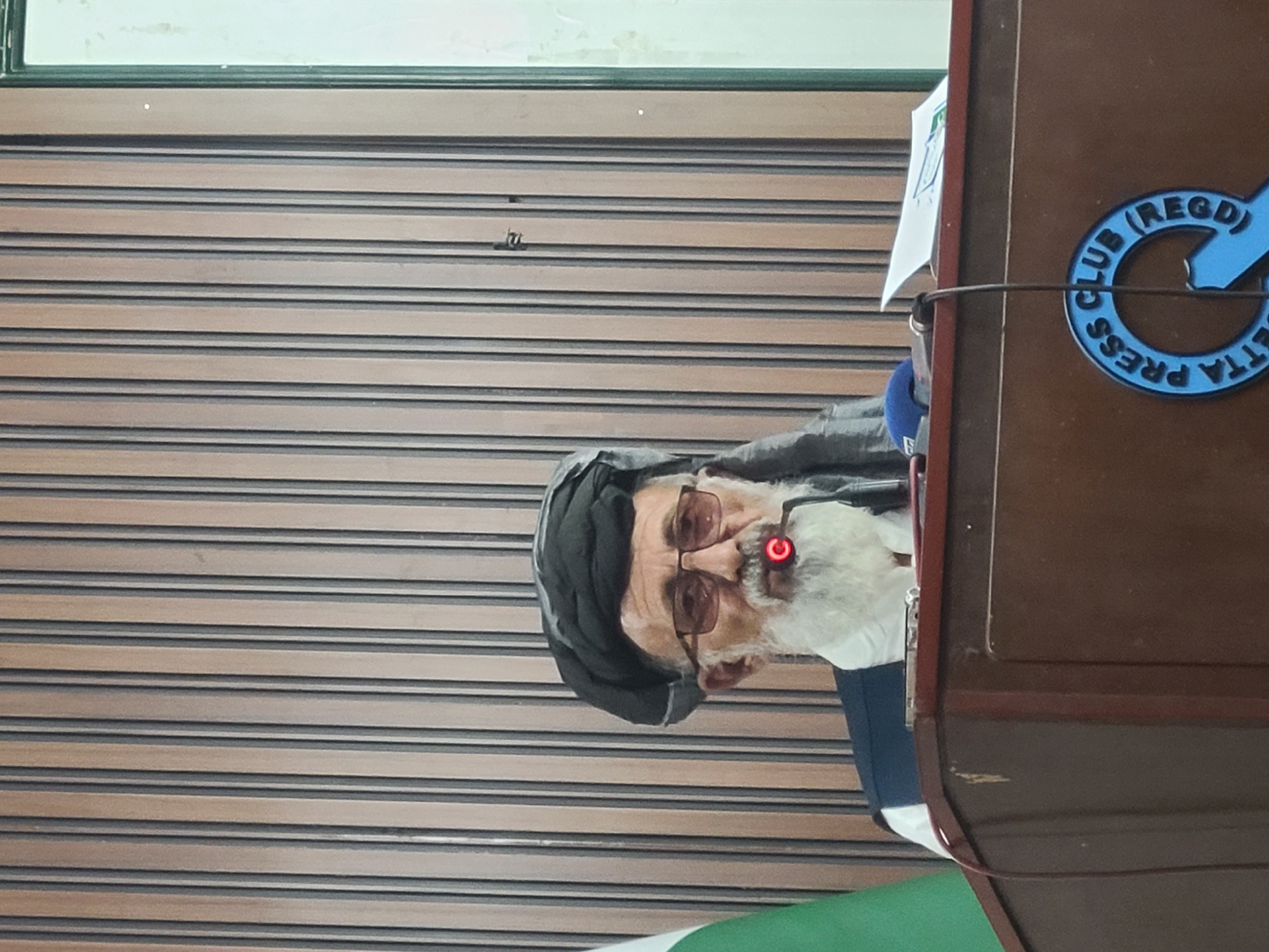 The image shows an elderly man with a white beard, wearing glasses and a black head covering, possibly a turban, standing behind a podium. A microphone is in front of him. The podium has a logo for the "Quetta Press Club (Regd)". The backdrop behind the man and podium is a closed, horizontally-striped shutter. The location is likely Quetta City Tehsil, Pakistan, due to the presence of the Quetta Press Club logo.