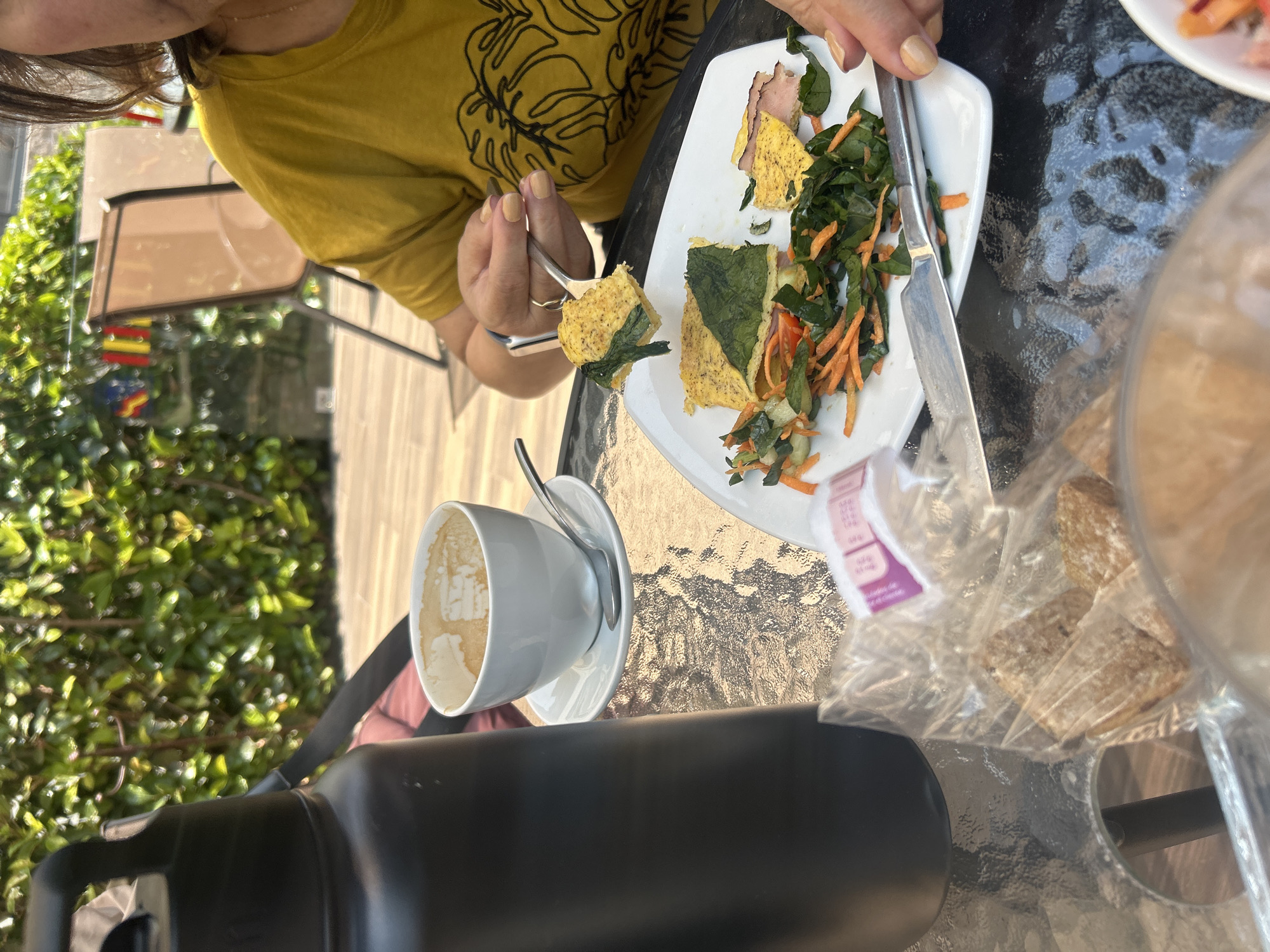 On a glass table, a person wearing a mustard-yellow shirt is eating from a white plate with a fork. The plate contains what appears to be an egg dish with spinach, carrots, and ham. A silver knife is on the side of the plate. Next to the plate is a half-empty white coffee cup on a saucer, a black water bottle, and bread in a plastic bag. The setting appears to be outdoors on a patio with green foliage visible in the background. The location is San Bernardino Tlaxcalancingo, Mexico.