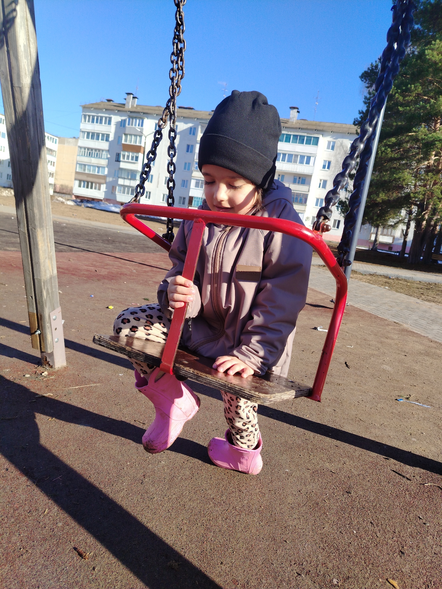 A young girl is sitting on a red and wood swing at a playground. She is wearing a black knit hat, a tan hooded jacket, animal print leggings, and pink rain boots. The swing is suspended by chains from a wooden frame. The playground surface is brown. In the background are apartment buildings, some with melting snow in front. Trees are also visible in the background. The image was taken in Noviy, Russia.